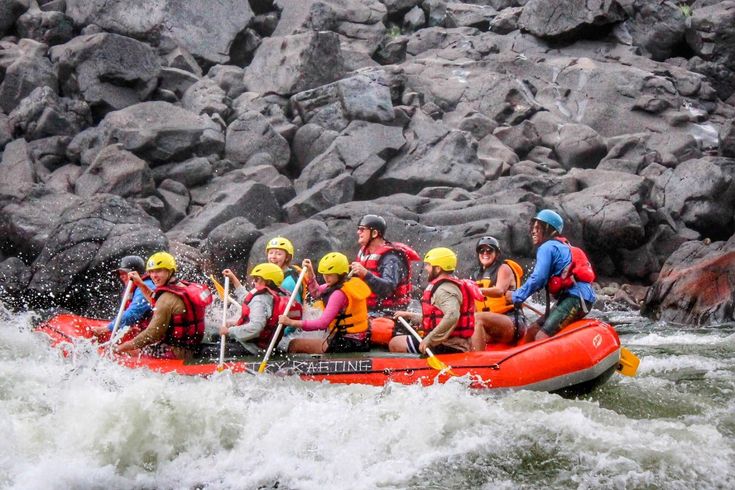 A raft with people navigating a river rapid, large grey rocks in the background.