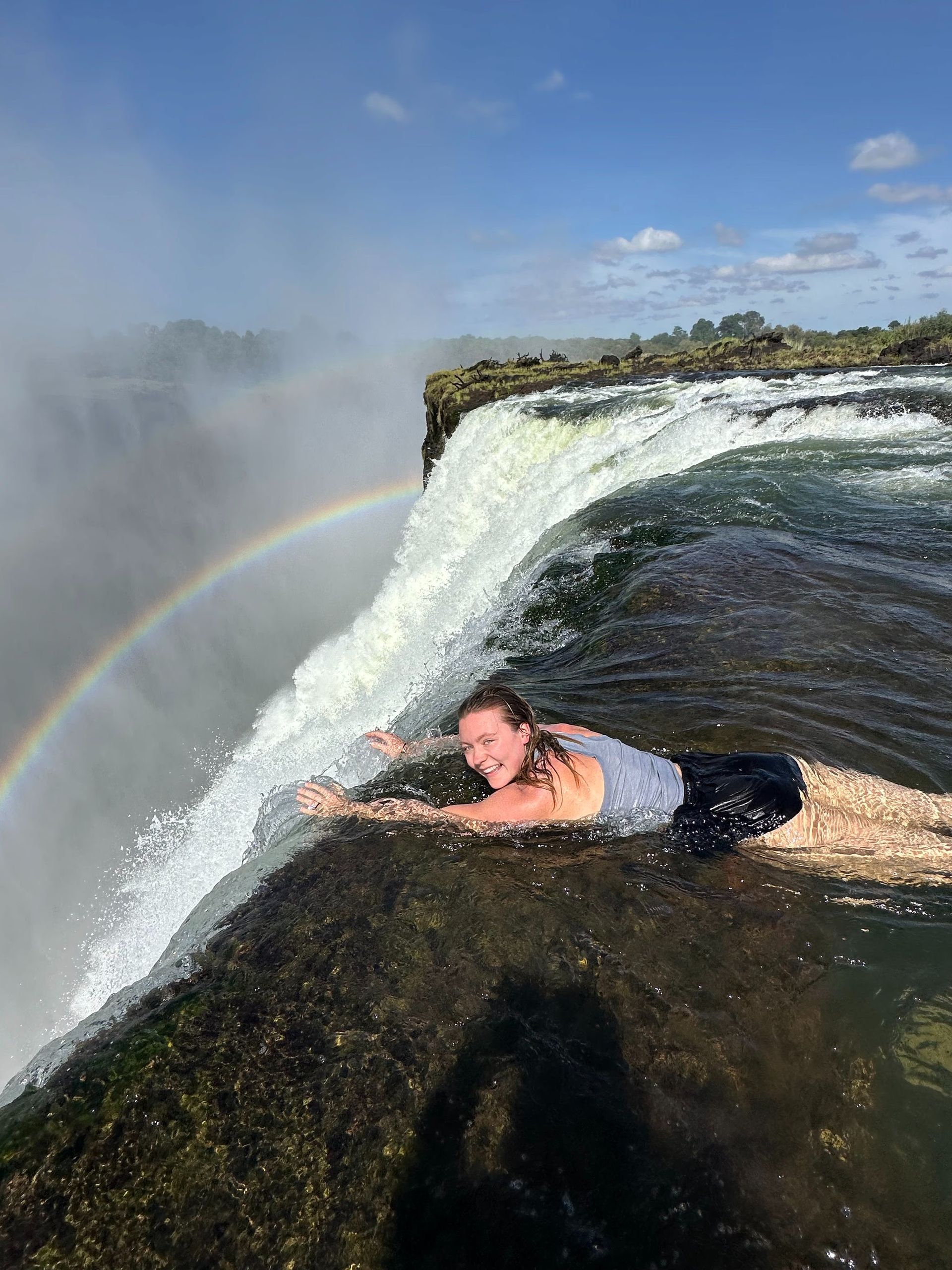 A woman is laying on the edge of a waterfall with a rainbow in the background.