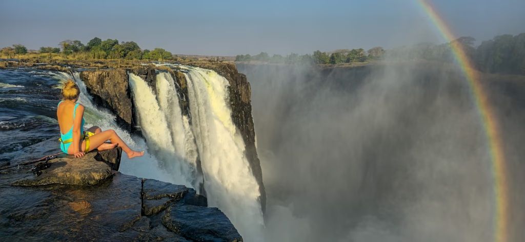 A woman is sitting on the edge of a waterfall with a rainbow in the background.