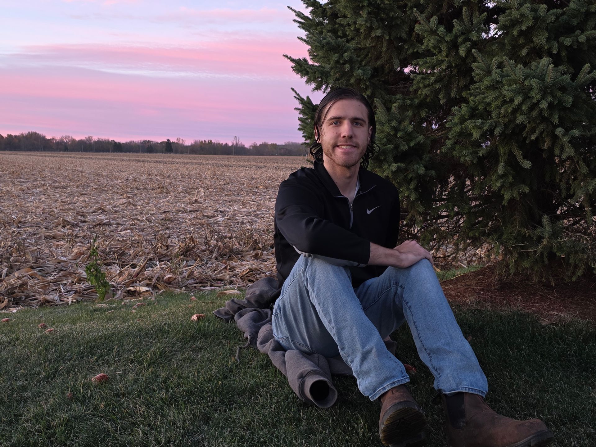 Man sitting on grass, smiling, in front of a field and pink sunset.
