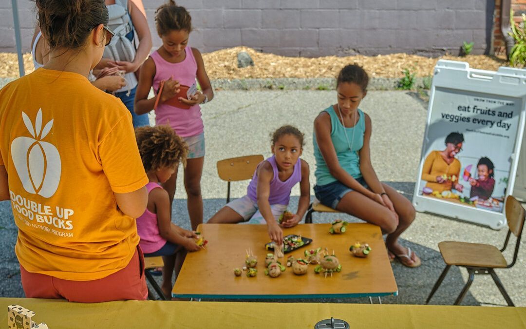 A woman in an orange shirt is standing next to a table with children sitting at it.