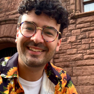 Man with curly hair and patterned glasses smiles in front of a brick building, wearing a sunflower shirt.