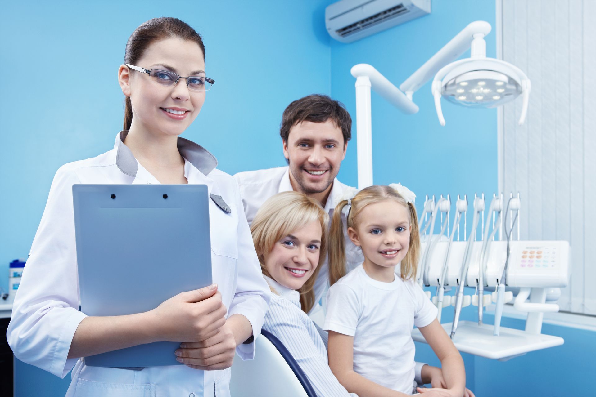 Female family dentist in white scrubs smiles at the camera next to family inside a dentist office.