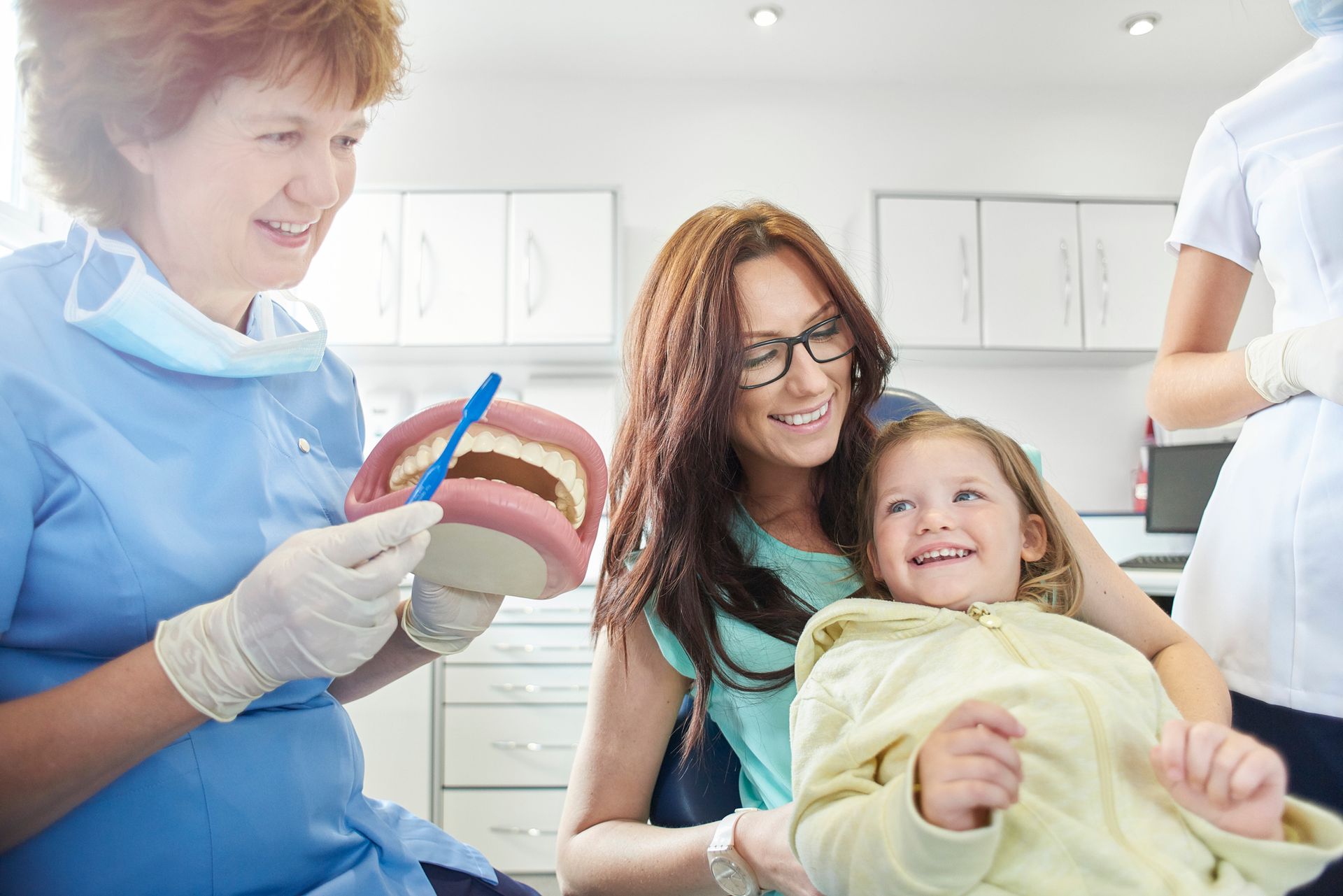 Female family dentist shows little girl and mother sitting in a chair how to brush a teeth model. Female family dentist shows little girl and mother sitting in a chair how to brush a teeth model.