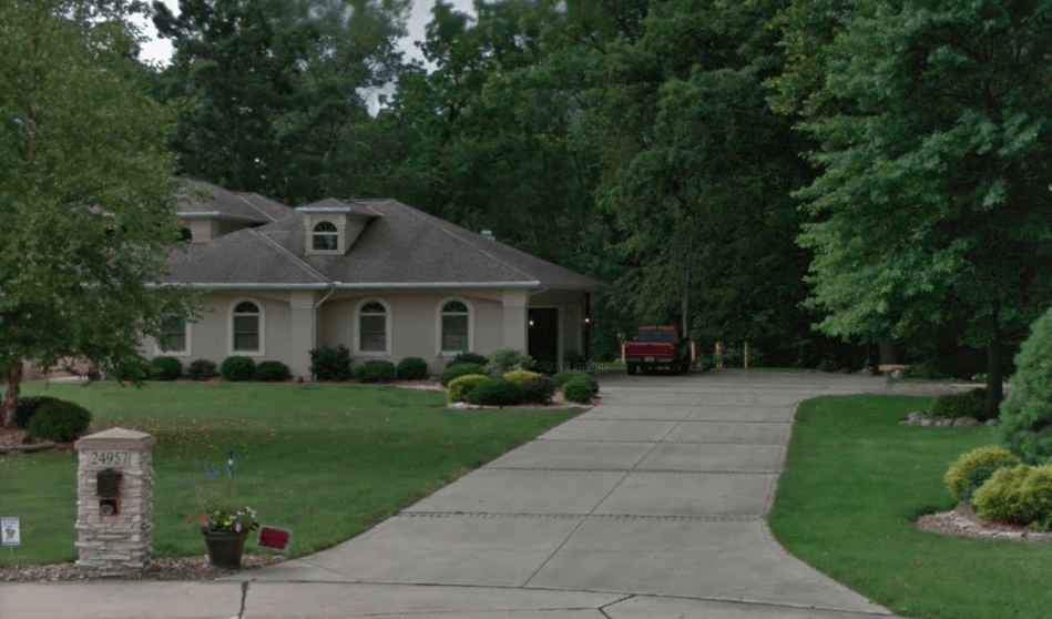 House with a long driveway, green lawn, and trees. Red truck parked in the carport.