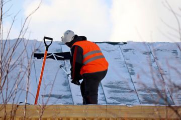 Un ouvrier du bâtiment portant un gilet orange et un casque de sécurité pelletant la neige d'un toit.
