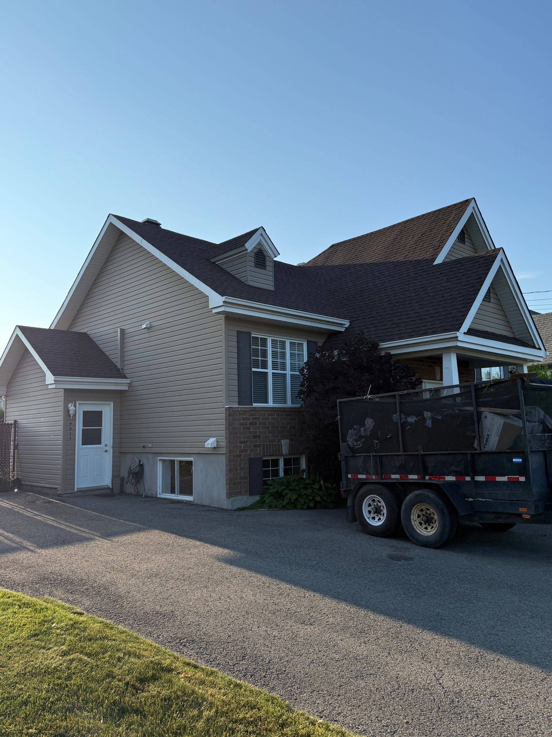 Maison beige avec toit marron foncé, allée en gravier et caravane. Ciel bleu.