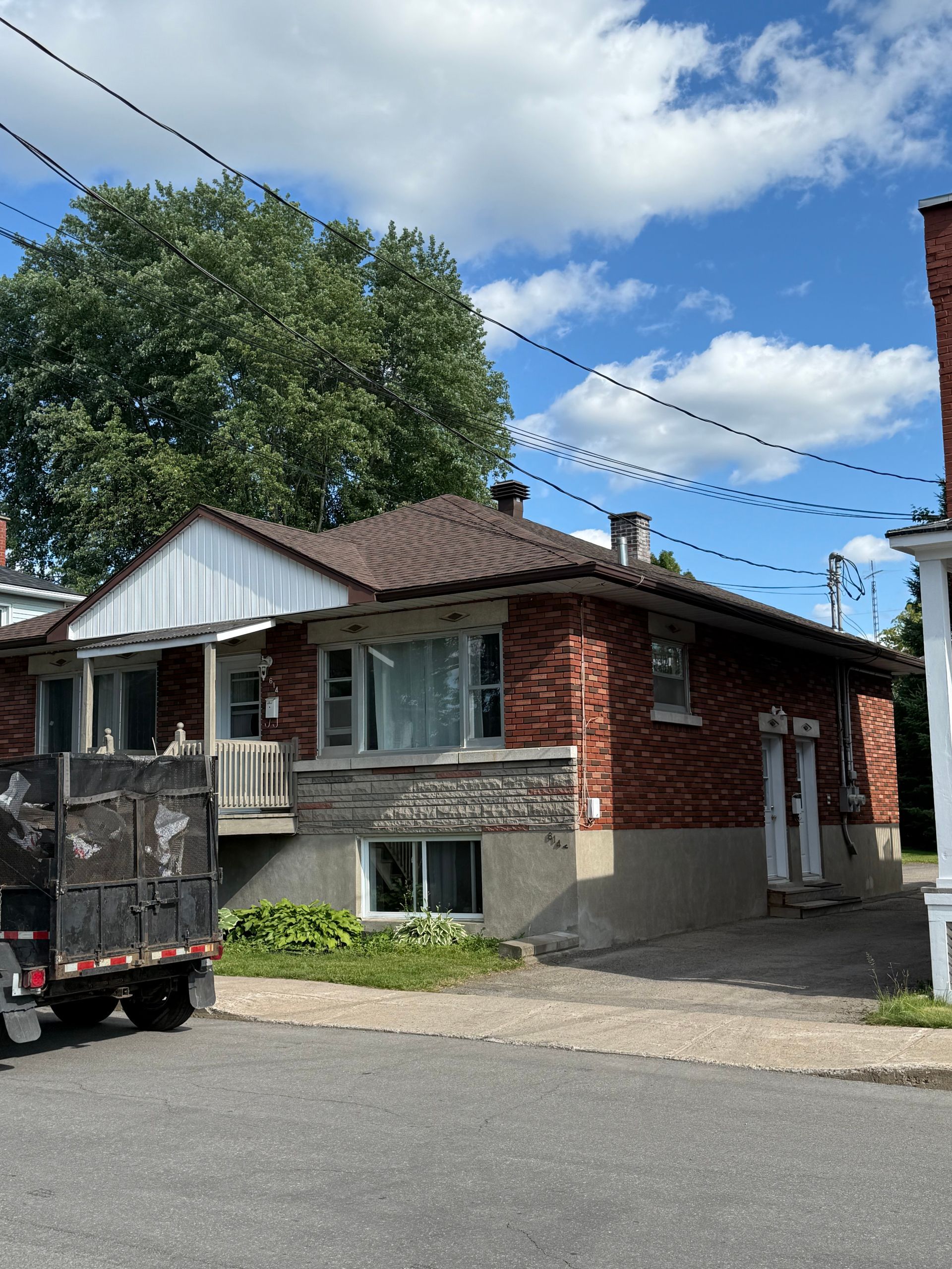 Maison en briques avec fondations apparentes. Pavé devant, verdure autour. Lignes électriques aériennes et ciel bleu.