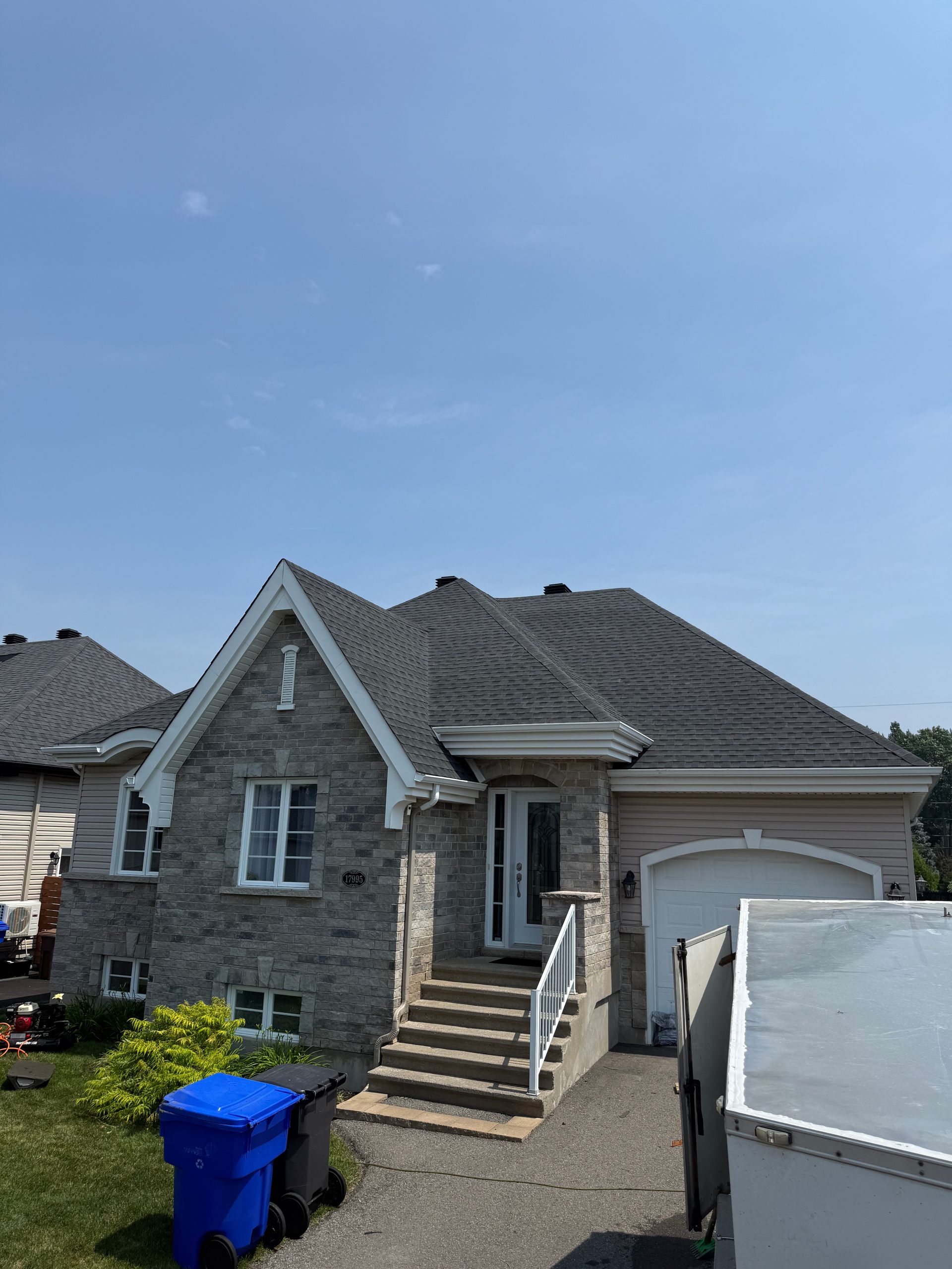 Maison en pierre grise avec toit noir et garnitures blanches, poubelle bleue devant, ciel bleu.