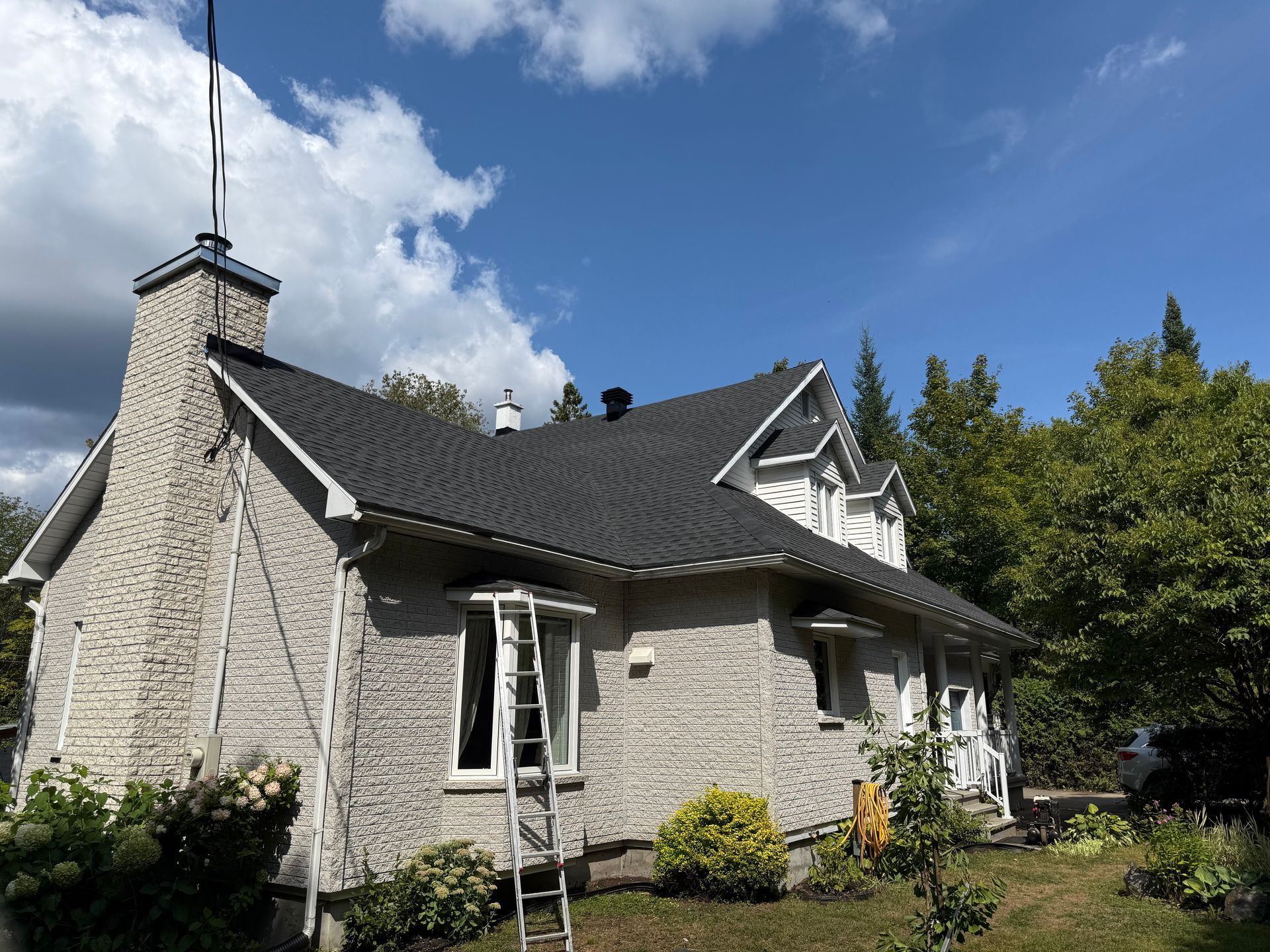 Maison en pierre grise avec un toit en bardeaux foncés, une échelle et un ciel clair.