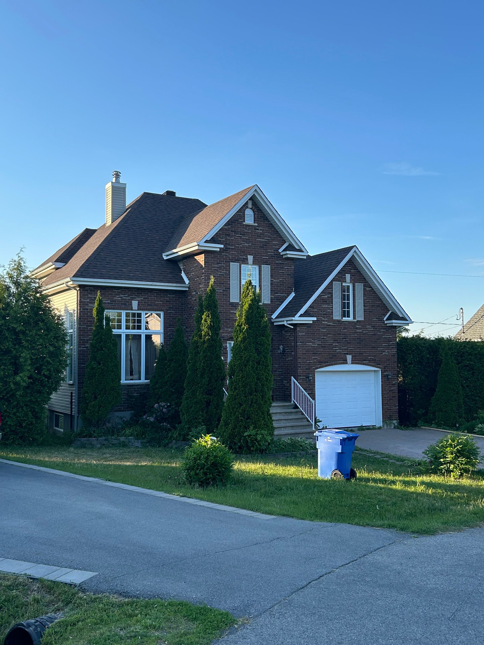 Maison en briques avec toit marron, garage blanc et conifères devant. Ciel bleu en arrière-plan.