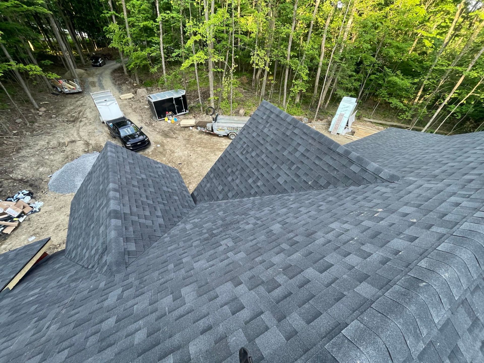 Vue depuis un toit avec des bardeaux gris ; chantier de construction avec des arbres, des véhicules et des fournitures visibles.