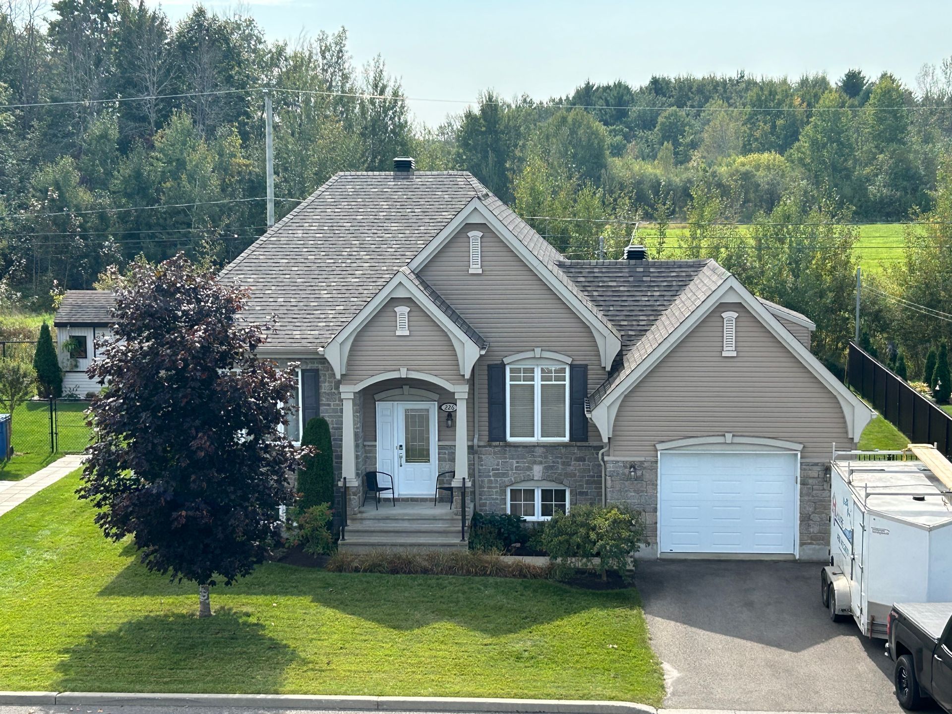 Maison beige et revêtue de pierre avec un toit gris, une porte de garage blanche et une pelouse verte.