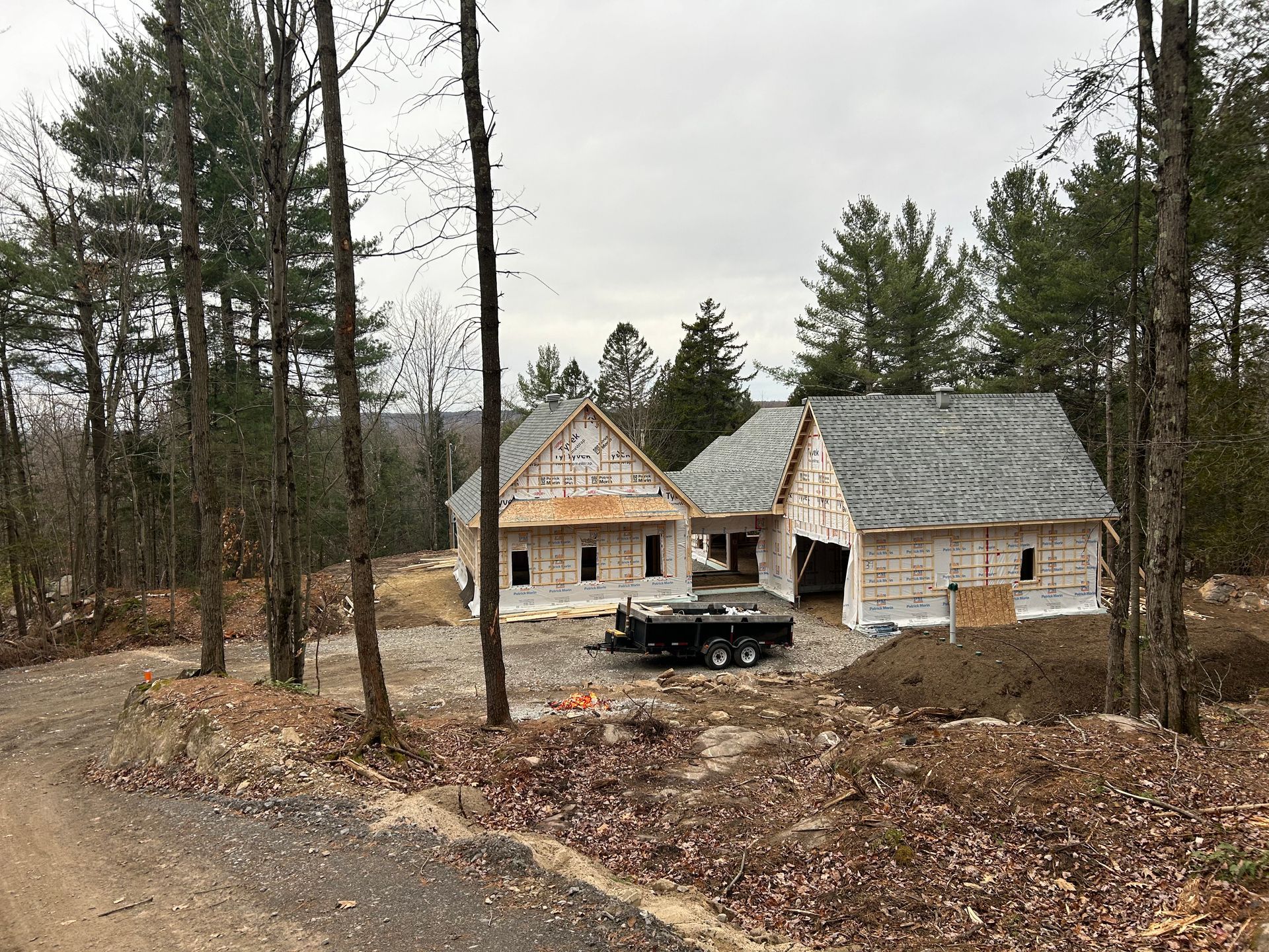 Maison en construction dans un secteur boisé avec charpente apparente et toiture temporaire; ciel gris couvert.