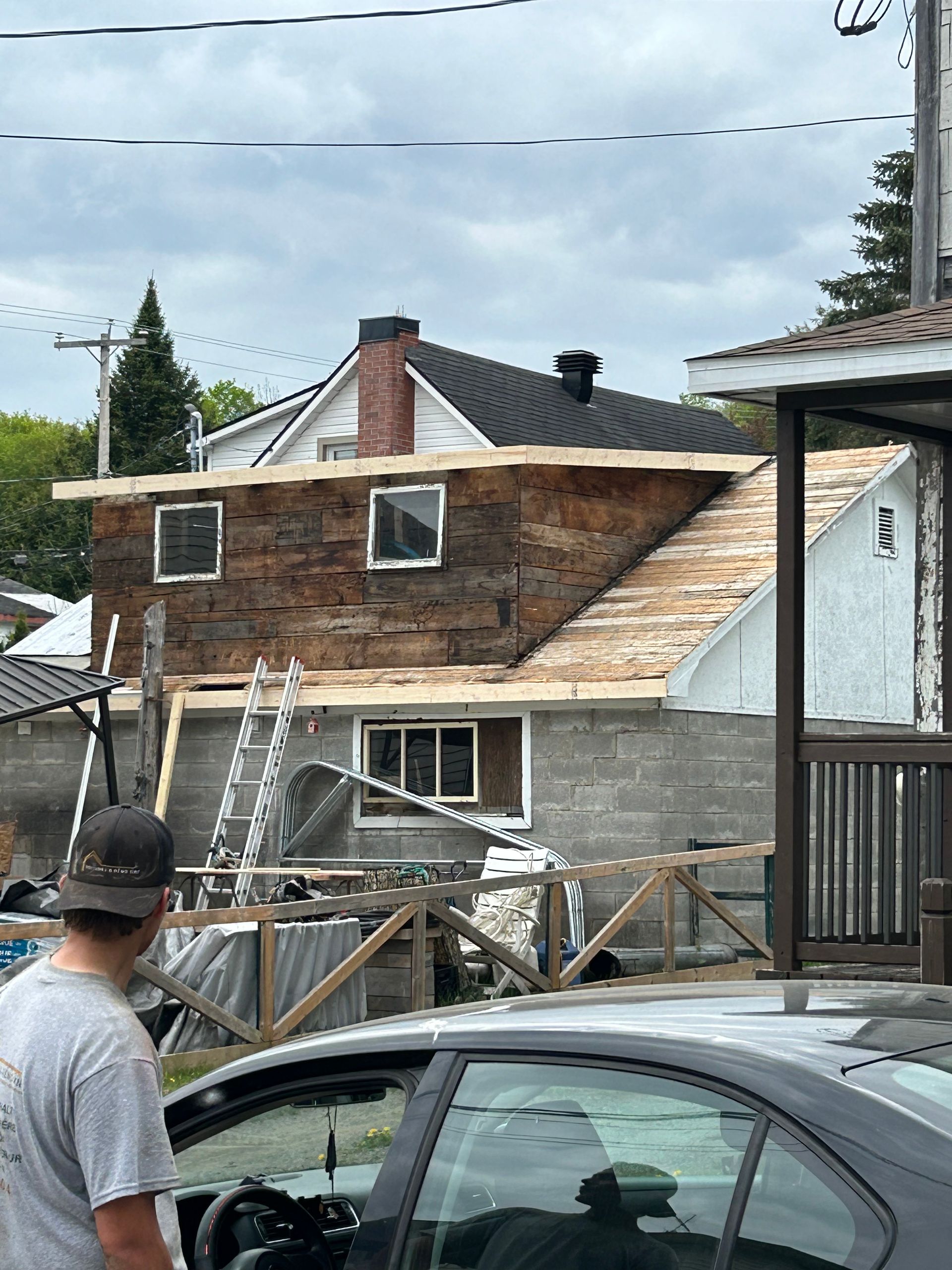 Construction d'une extension de maison à deux étages, revêtement en bois brun, parpaing gris, homme regardant, voiture au premier plan.