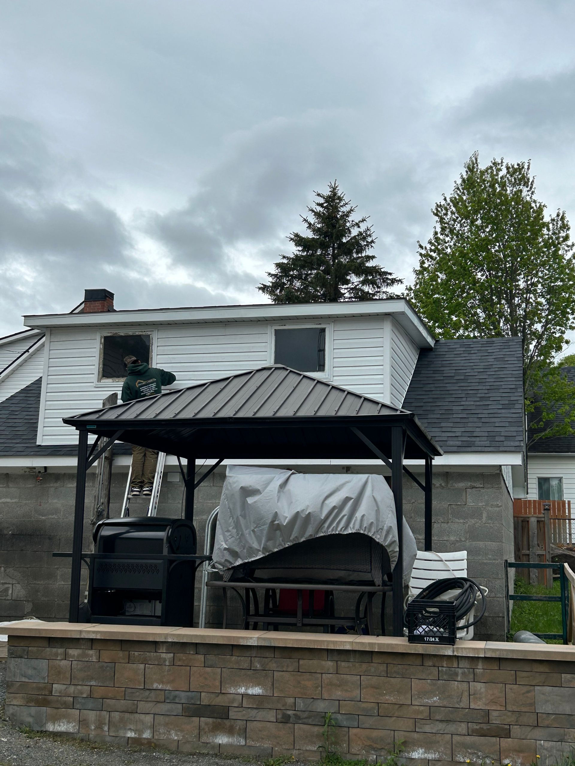 Un homme sur une échelle travaille sur une maison avec un nouveau revêtement. Un belvédère sombre devant, un ciel couvert.
