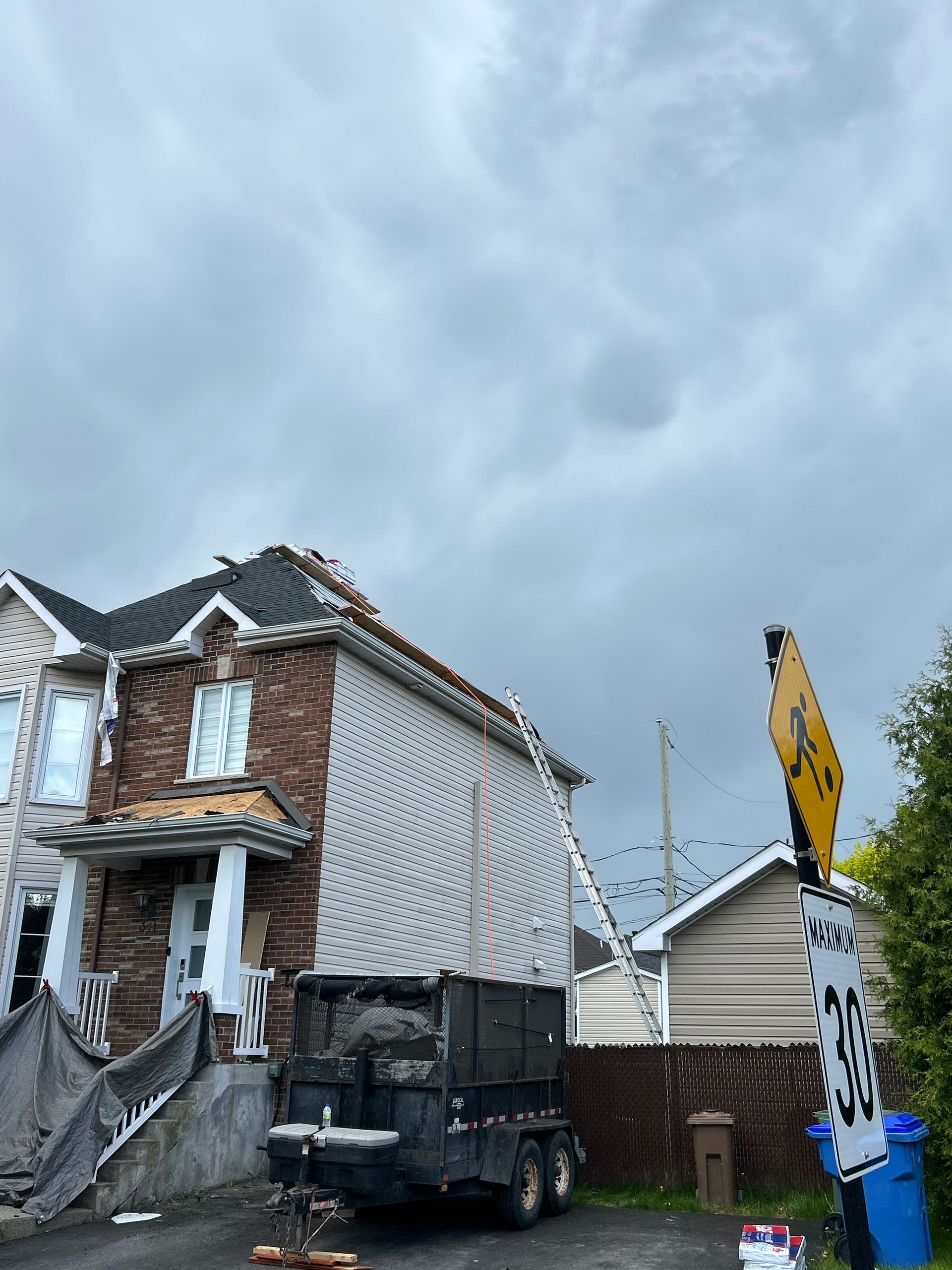 Maison avec revêtement manquant, une échelle et une benne à ordures sous un ciel nuageux.