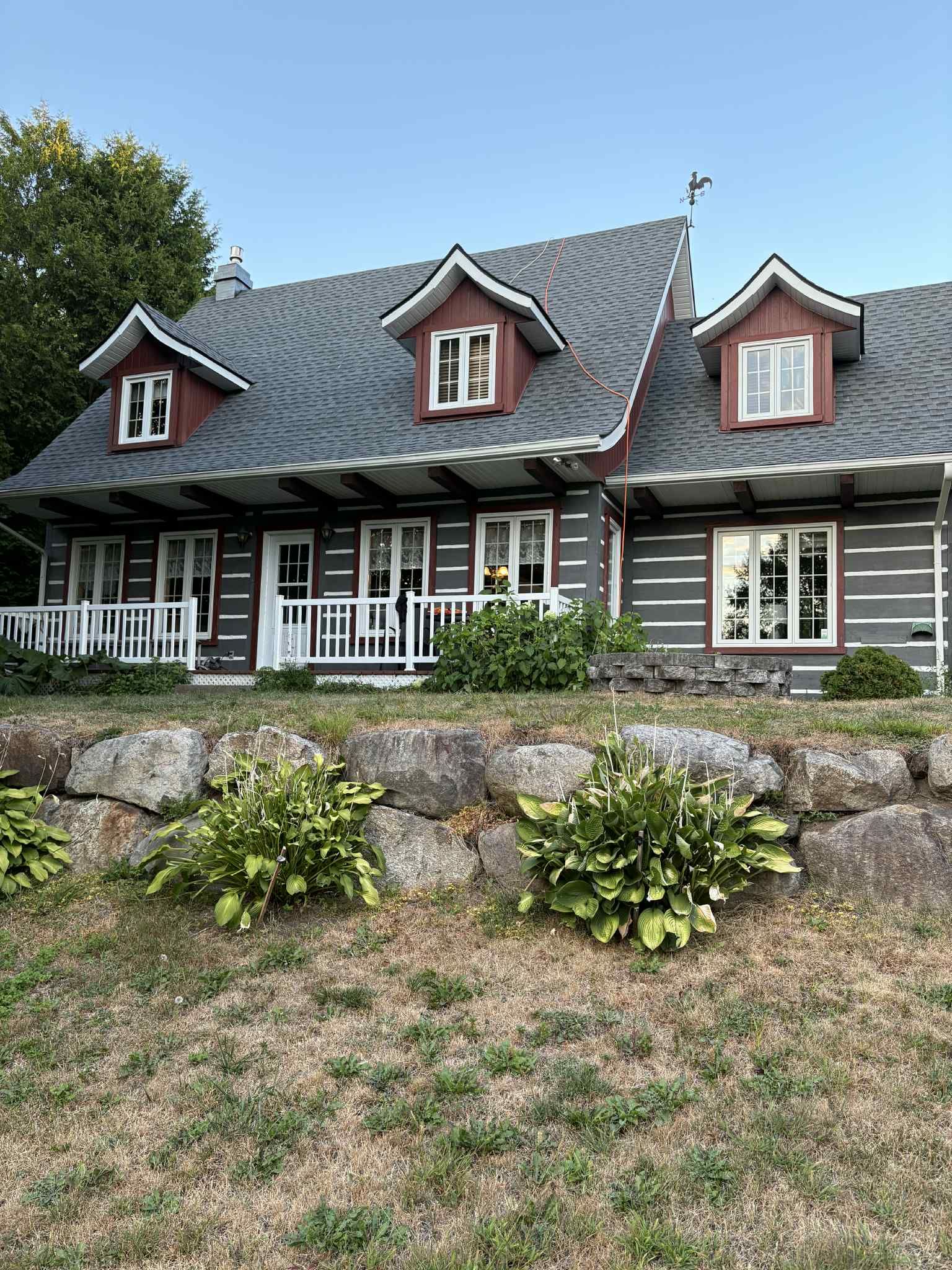 Cabane en rondins avec rondins gris, garnitures rouges et accents blancs, nichée derrière un mur de pierre et de la verdure.