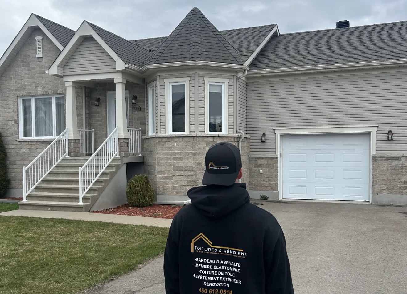 Personne portant un sweat à capuche noir face à une maison en briques grises avec une porte de garage blanche et des marches.