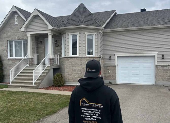 Une personne fait face à une maison claire avec garage. Elle porte un sweat à capuche et un chapeau noirs. Ciel nuageux, herbe verte.