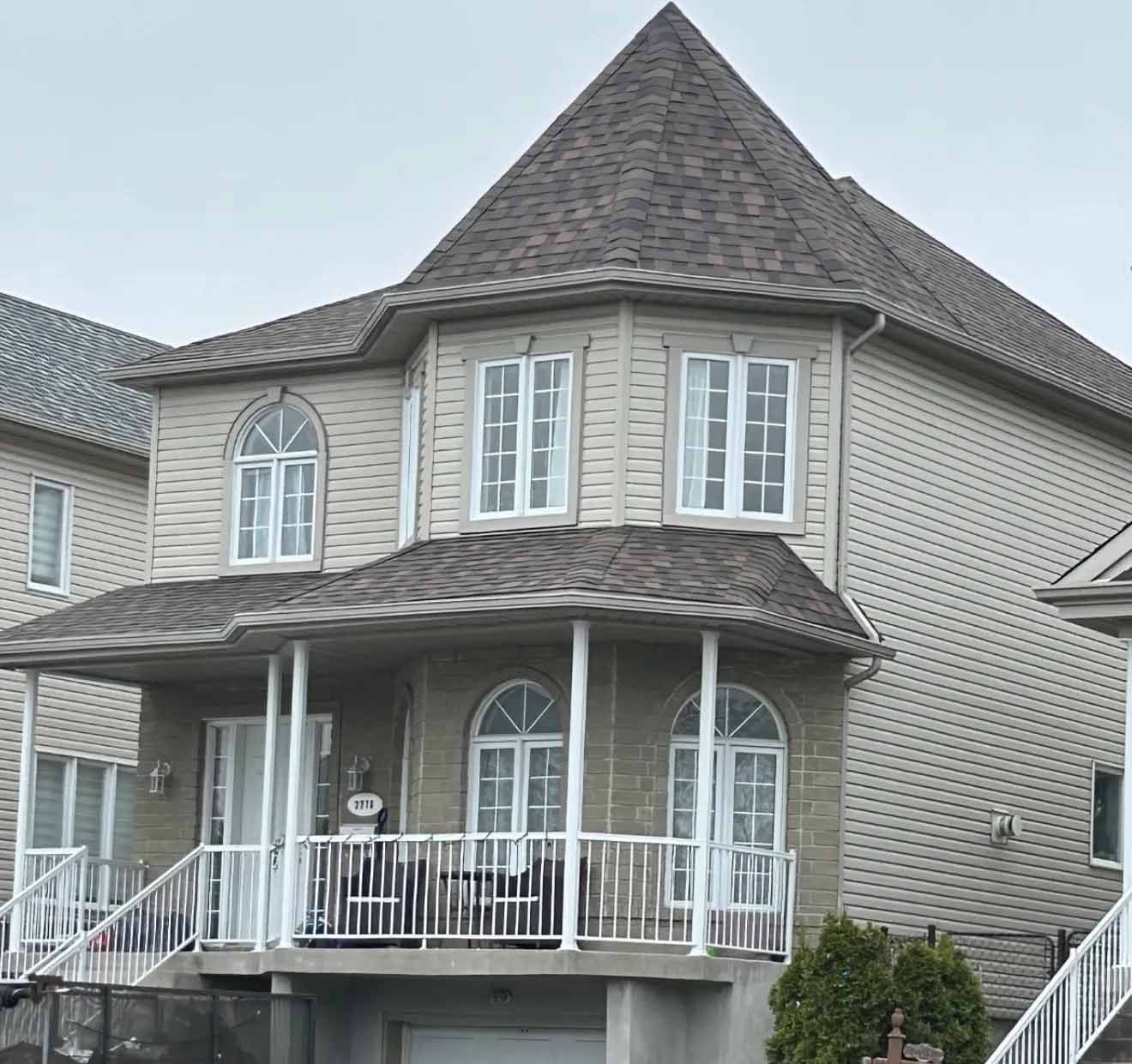 Maison beige à deux étages avec toit marron, balcon, fenêtres cintrées et balustrades blanches.