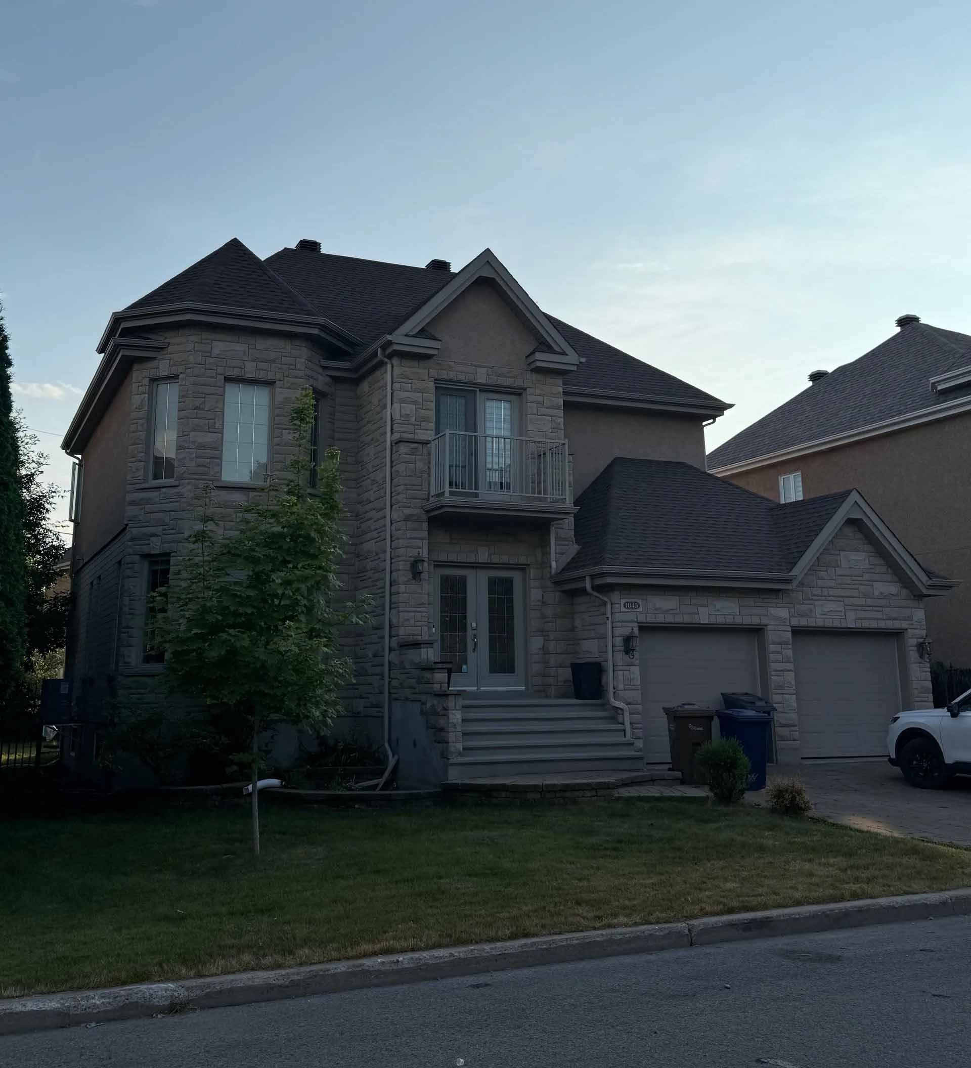 Maison en pierre à deux étages avec un balcon au-dessus de la porte d'entrée, un garage pour deux voitures et un petit arbre dans la cour.