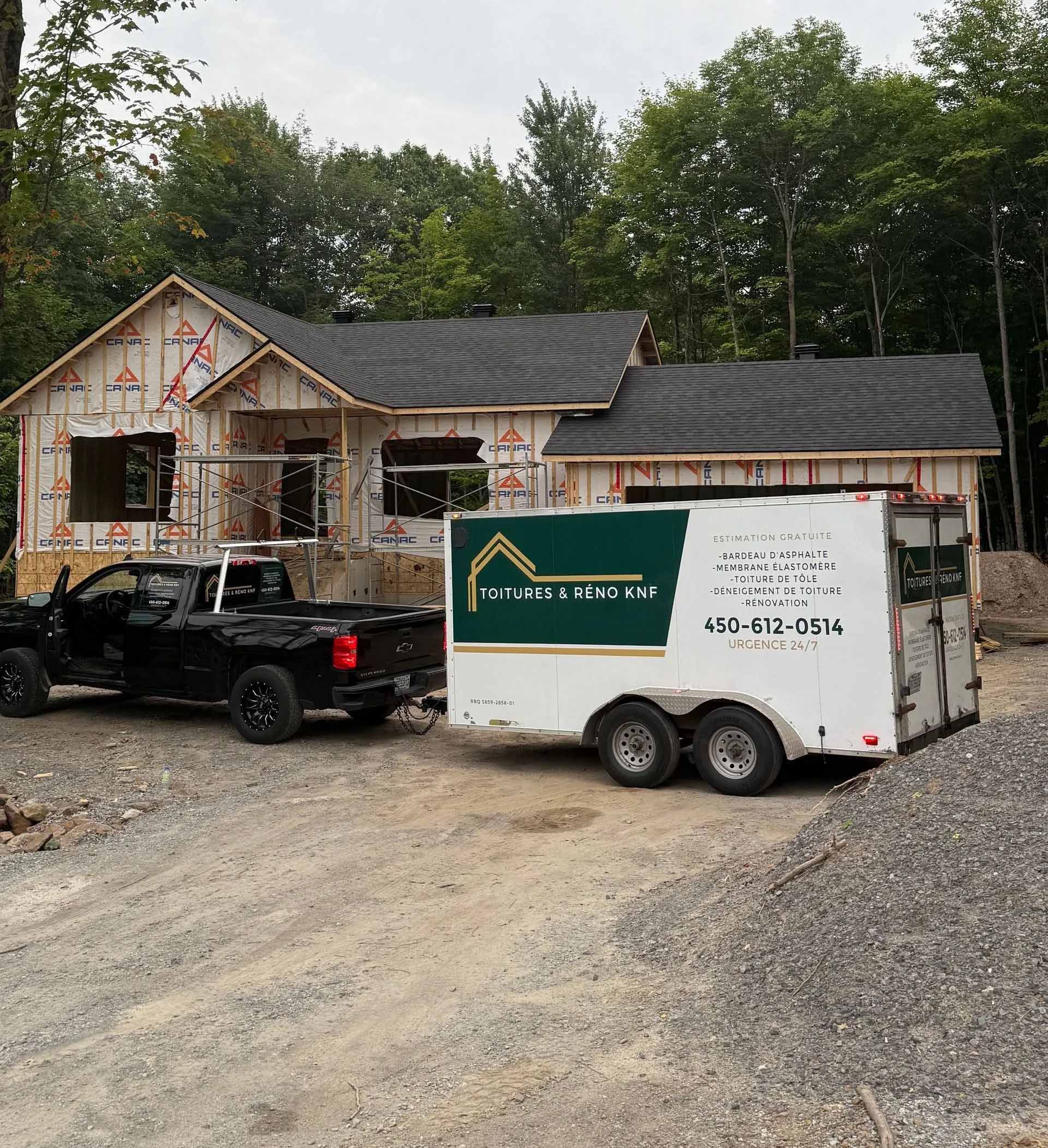 Camion noir tractant une remorque sur un chantier de construction d'une maison ; bois en arrière-plan.
