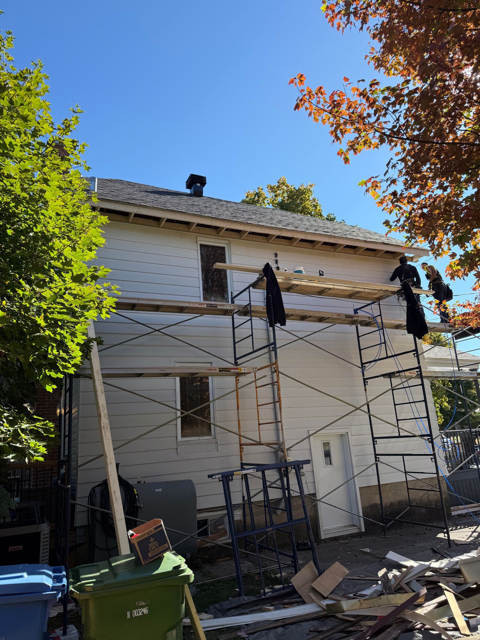 Des ouvriers sur un échafaudage revêtent une maison blanche avec un ciel bleu et un feuillage d'automne.
