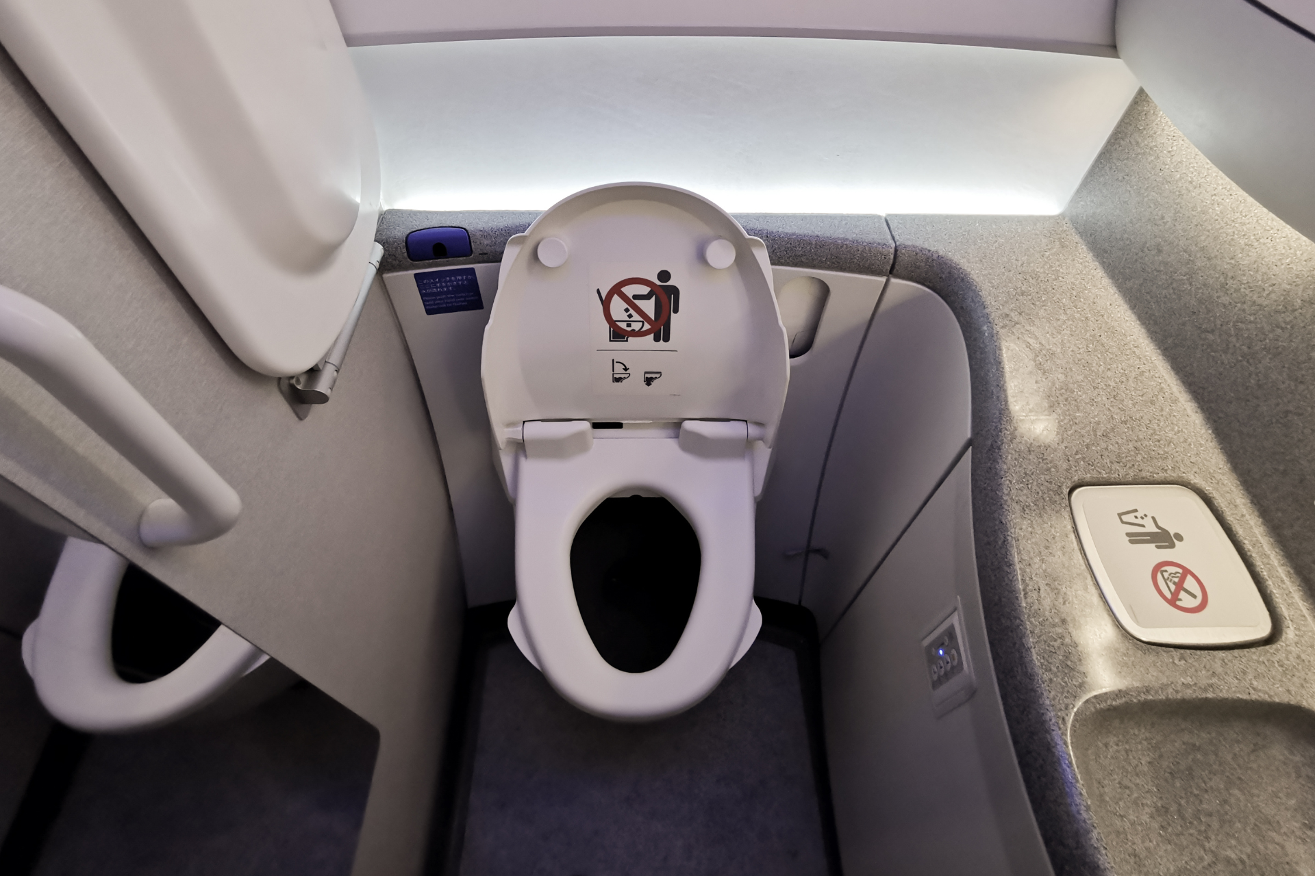 Airplane lavatory with open toilet lid, gray walls, and folded safety instructions beside the seat