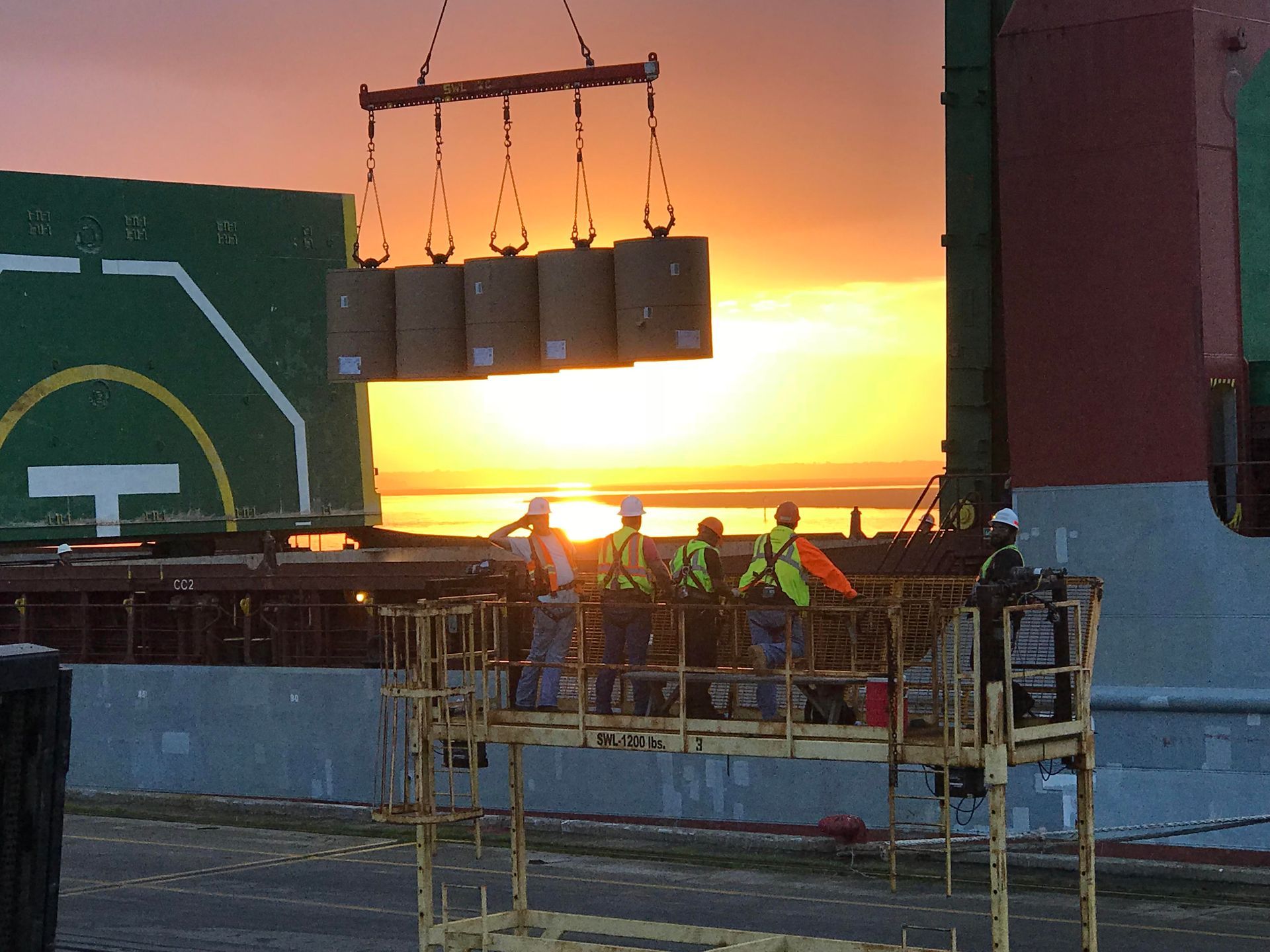 Workers on a ship deck at sunset lifting cargo with a crane, with the ocean glowing behind them.