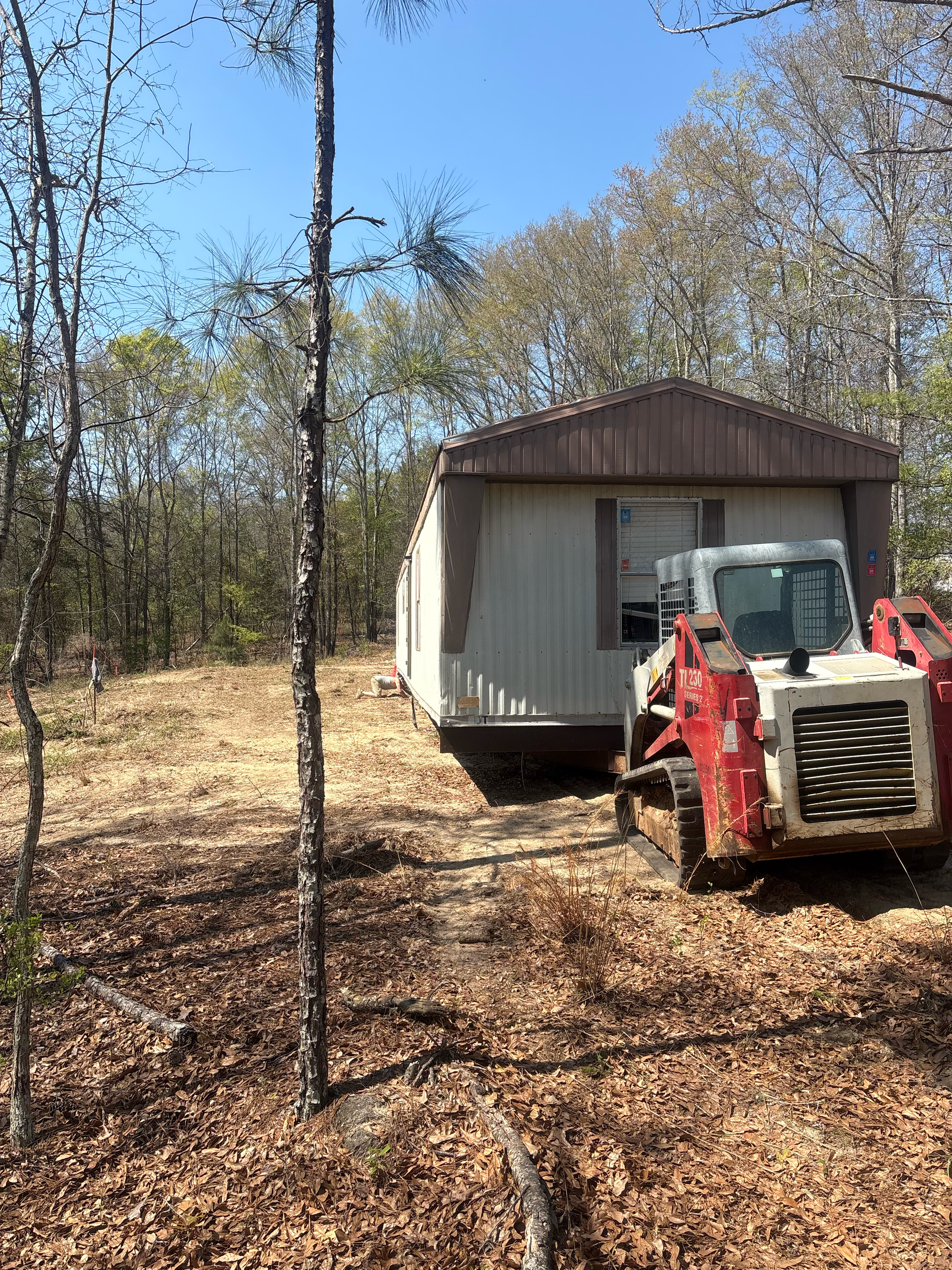 A red skid steer loader parked next to a mobile home in a wooded, sunlit area.
