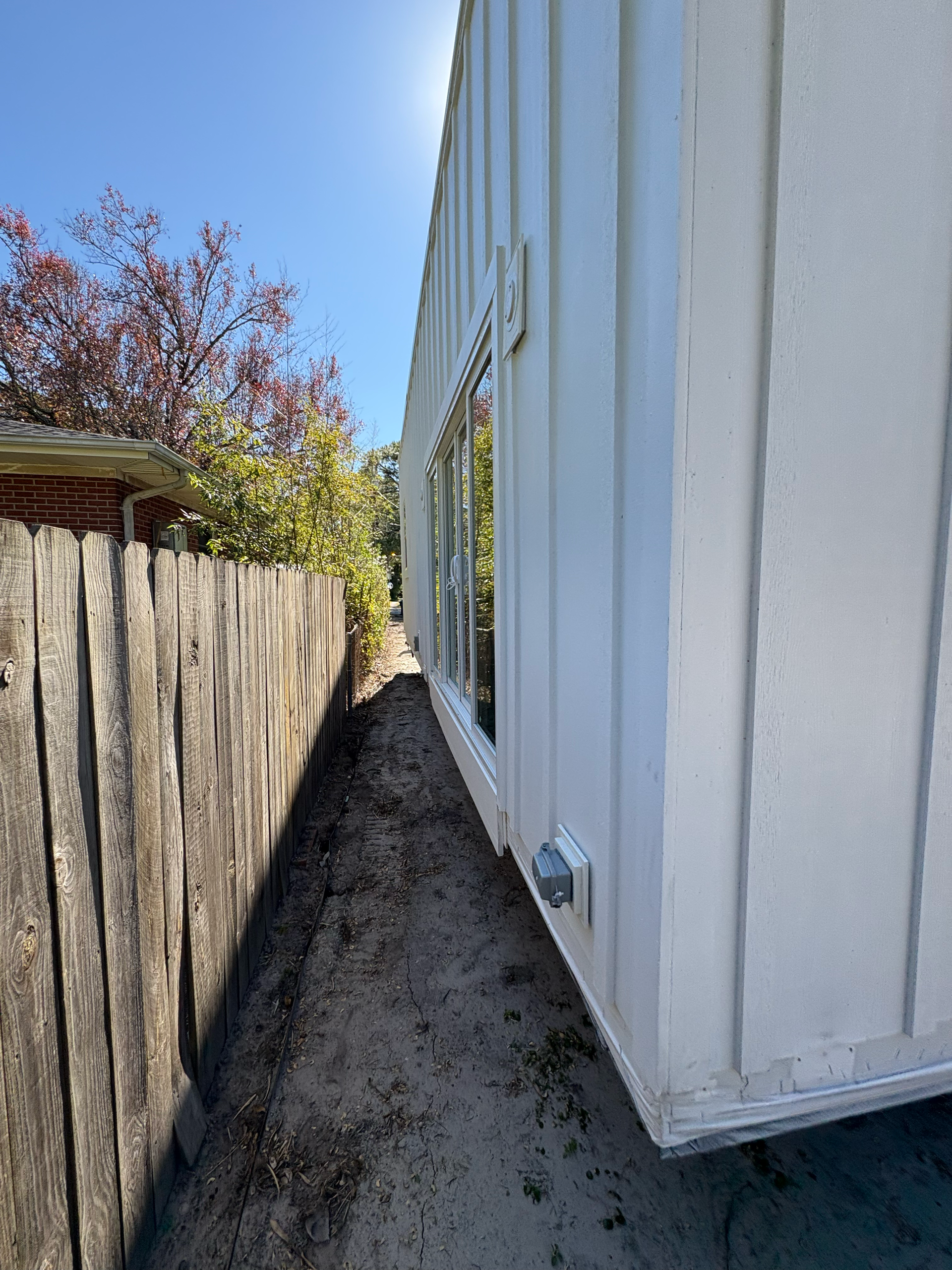 Narrow, dirt-filled side yard between a white house with vertical siding and a wooden fence.