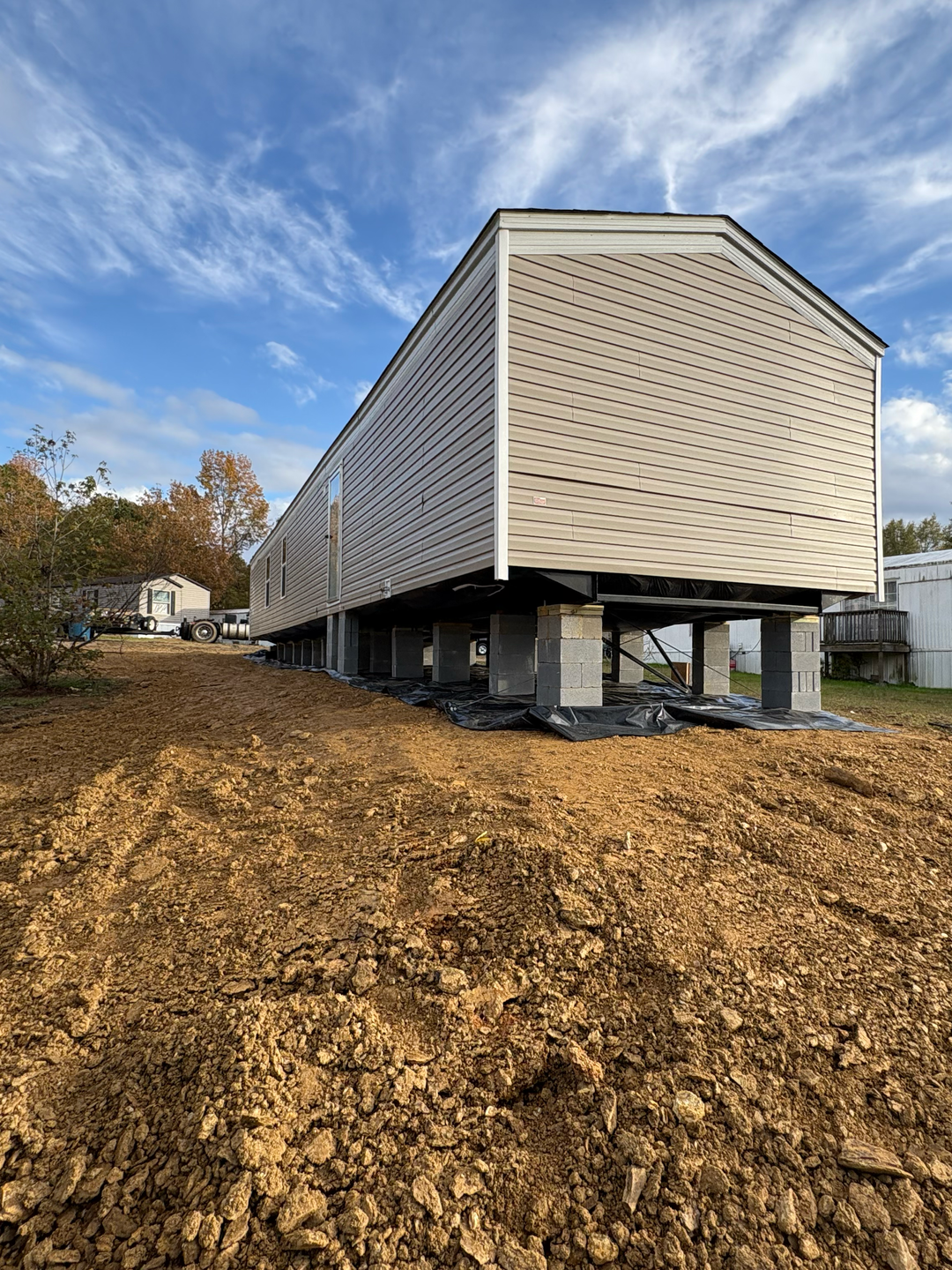 A long, beige manufactured home elevated on concrete block piers sits on a graded dirt lot under a blue sky with clouds.