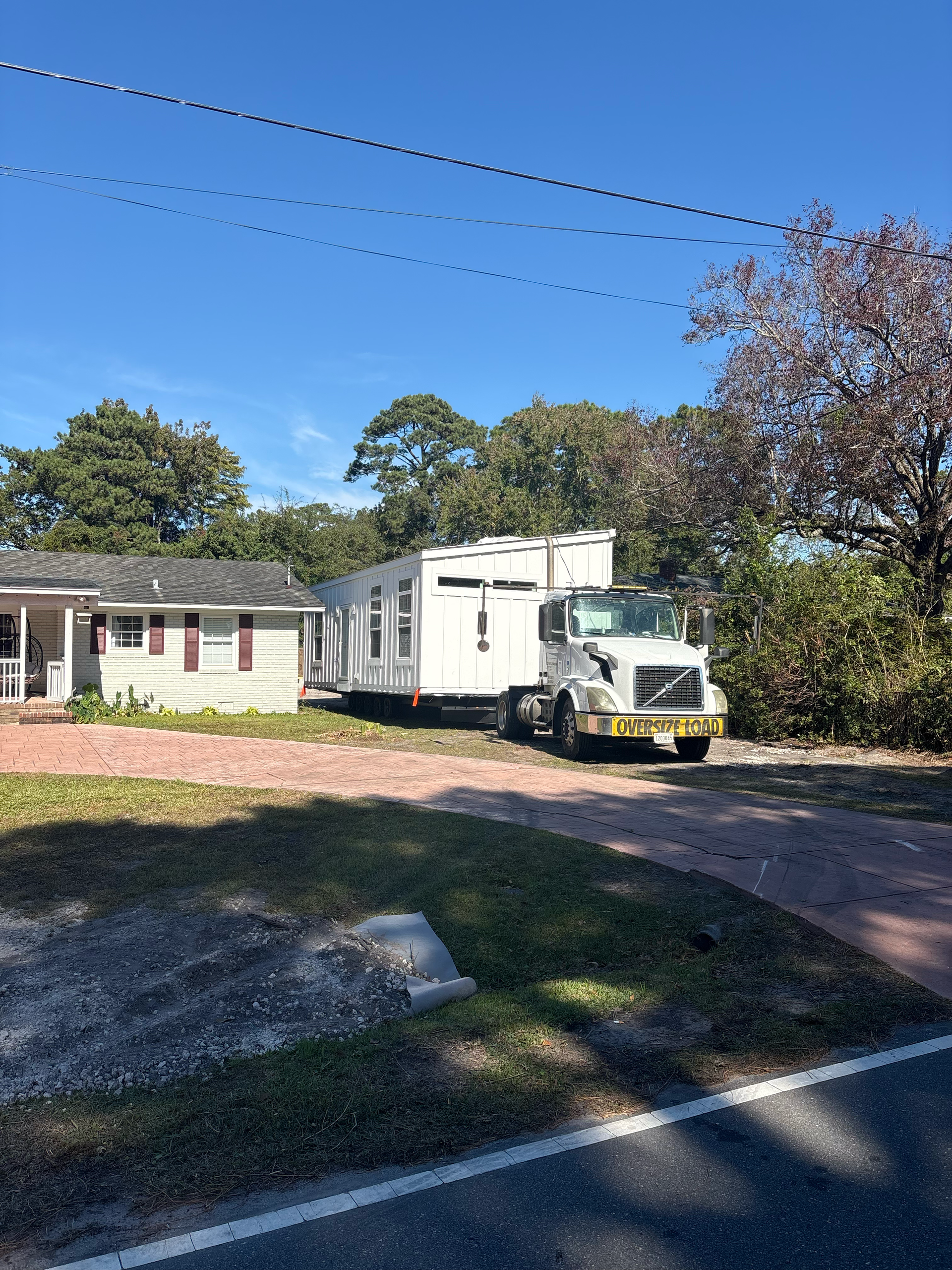 A white semi-truck hauls a white mobile home unit along a driveway near a small house on a sunny day.