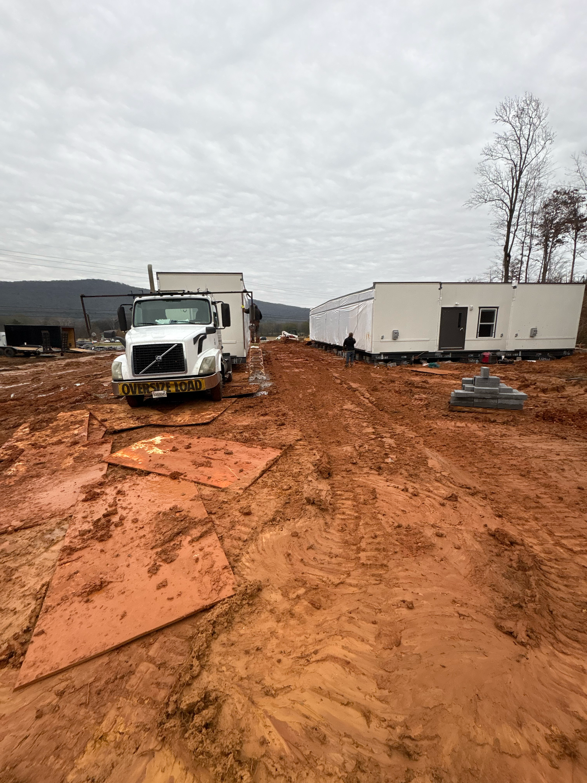 A white semi-truck pulls a trailer through a muddy construction site toward a pre-fabricated building under a gray sky.