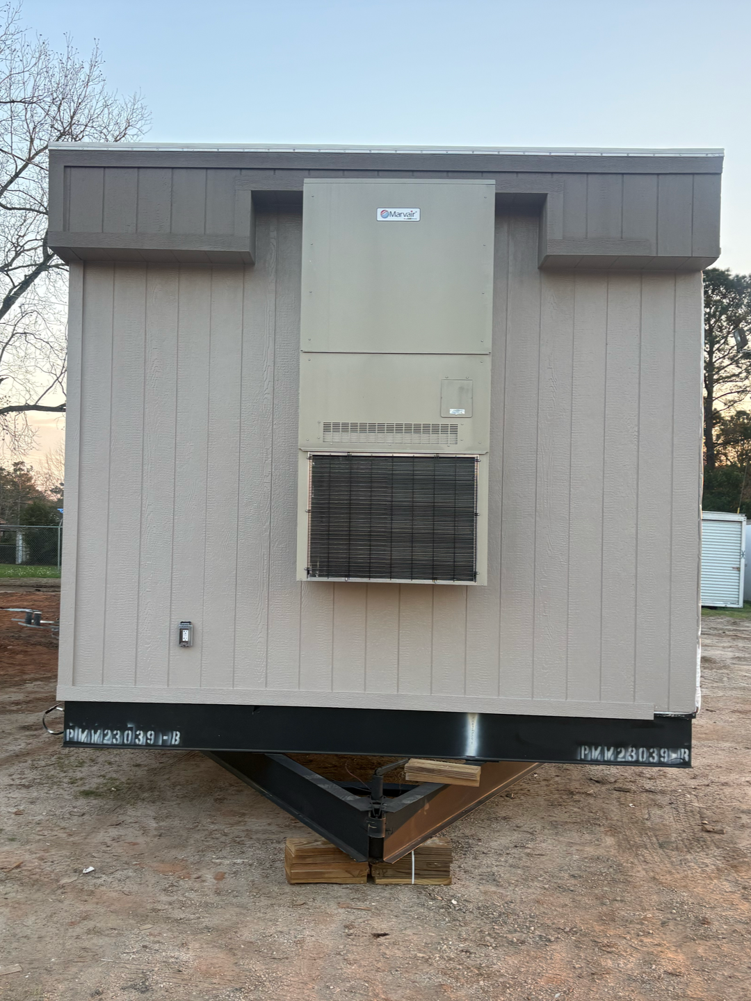 A beige mobile home exterior with a mounted HVAC unit, resting on wooden blocks in a dirt lot.