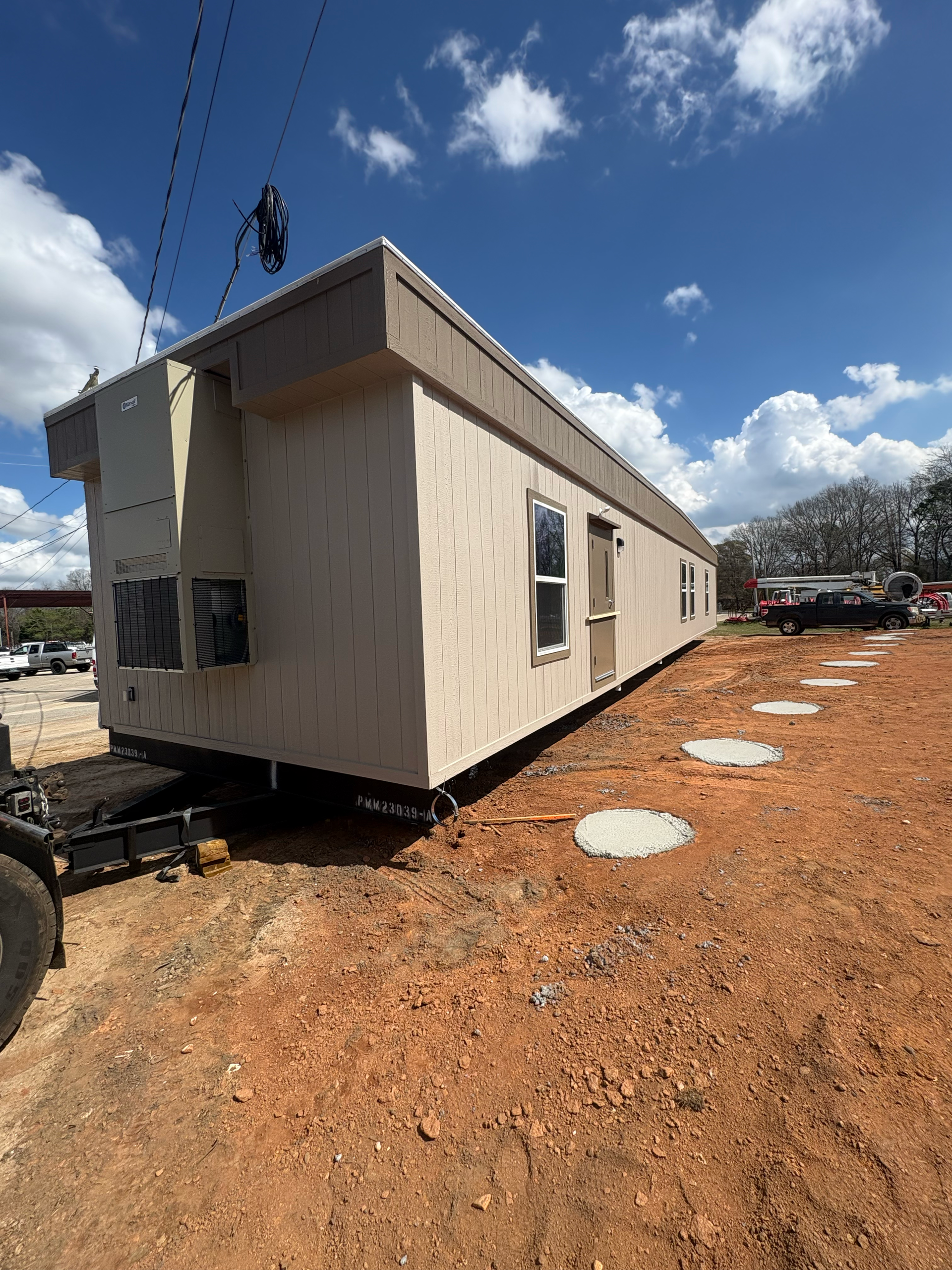 A tan manufactured home on a trailer frame sits on a red dirt lot next to circular concrete foundation footings.