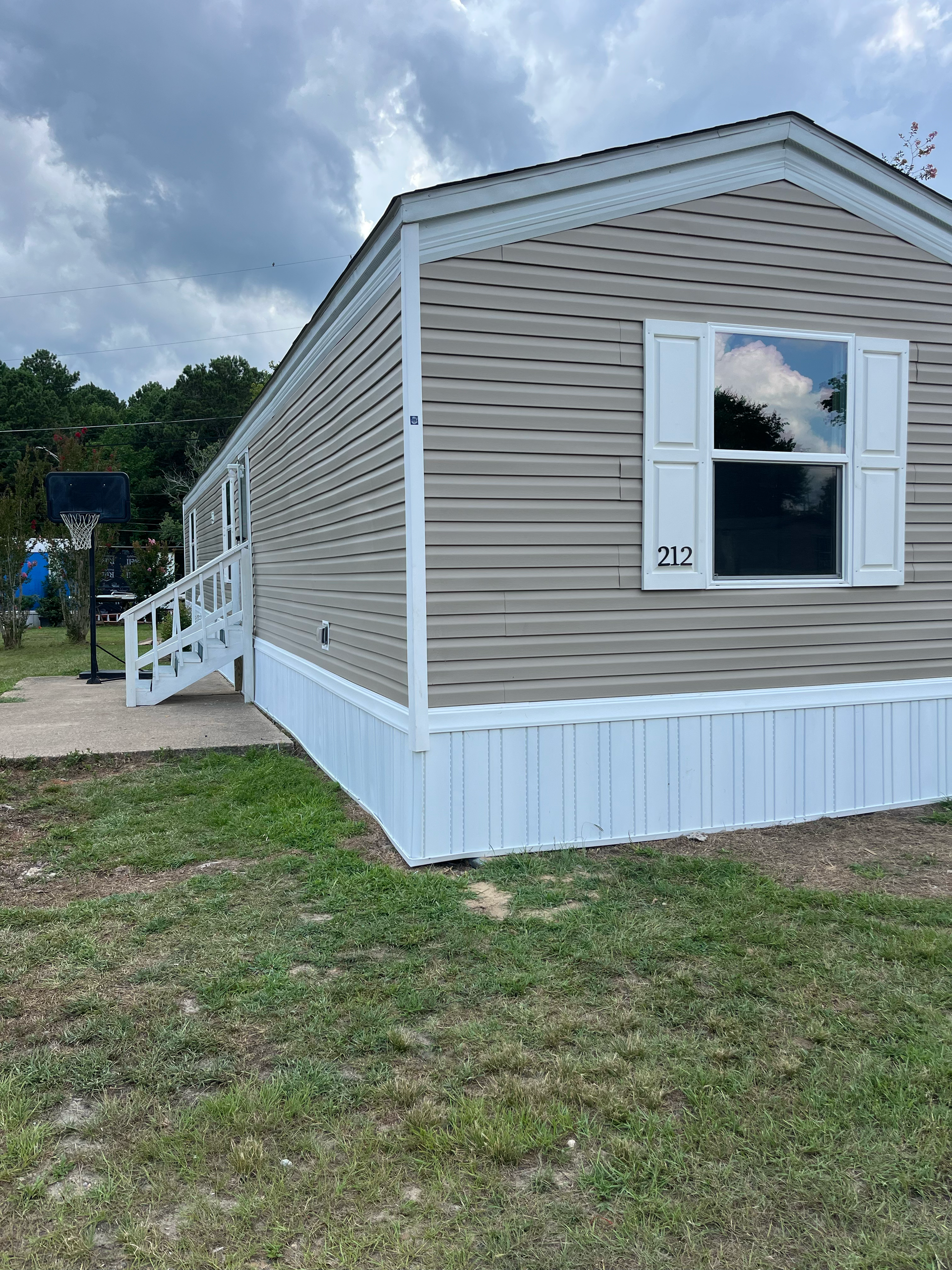 Tan mobile home with white vertical skirting, white shutters on a single window, and stairs leading to a side entry.