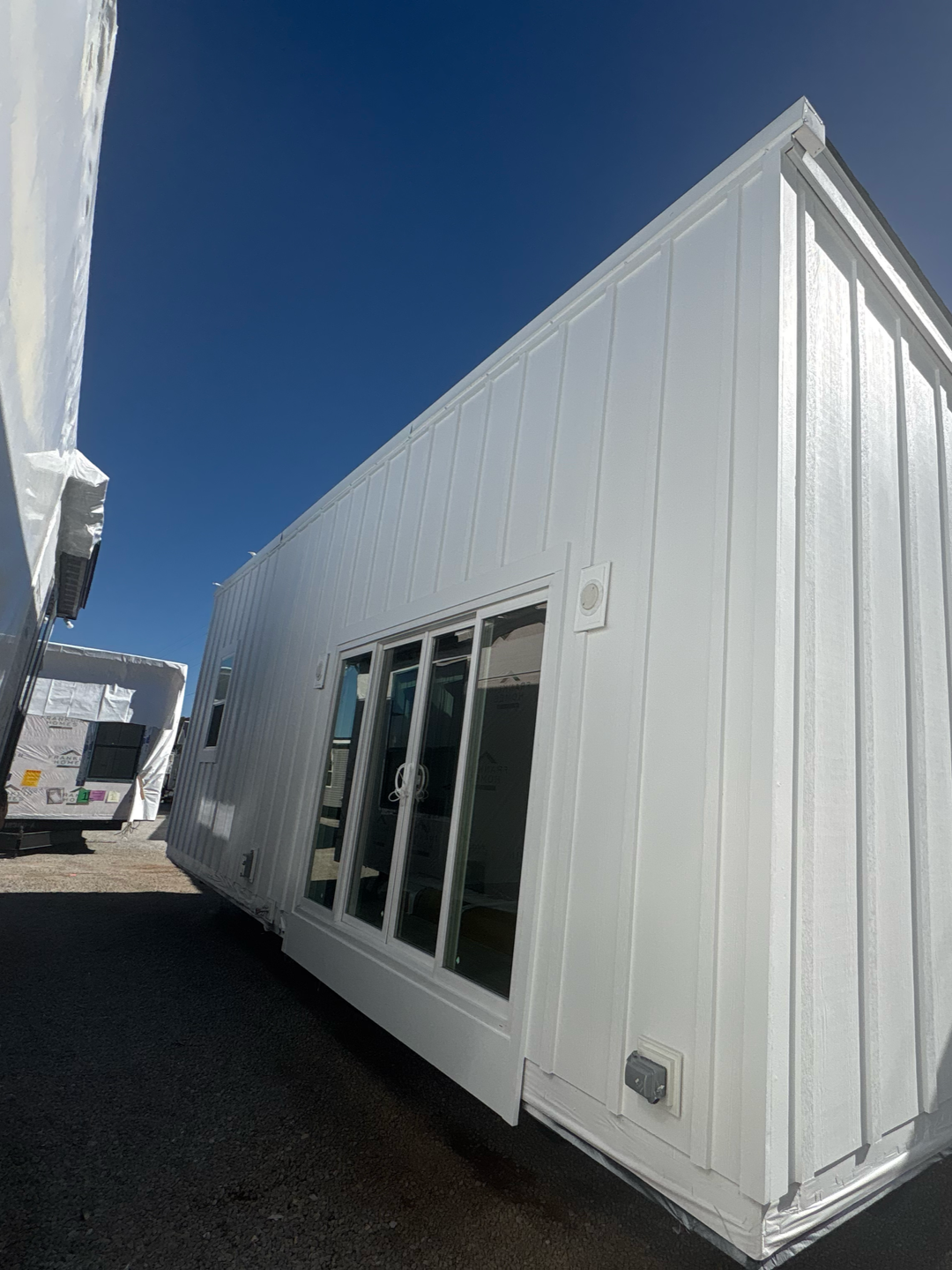 Low-angle view of a white, modern tiny home with vertical siding and large sliding glass doors in a gravel lot.
