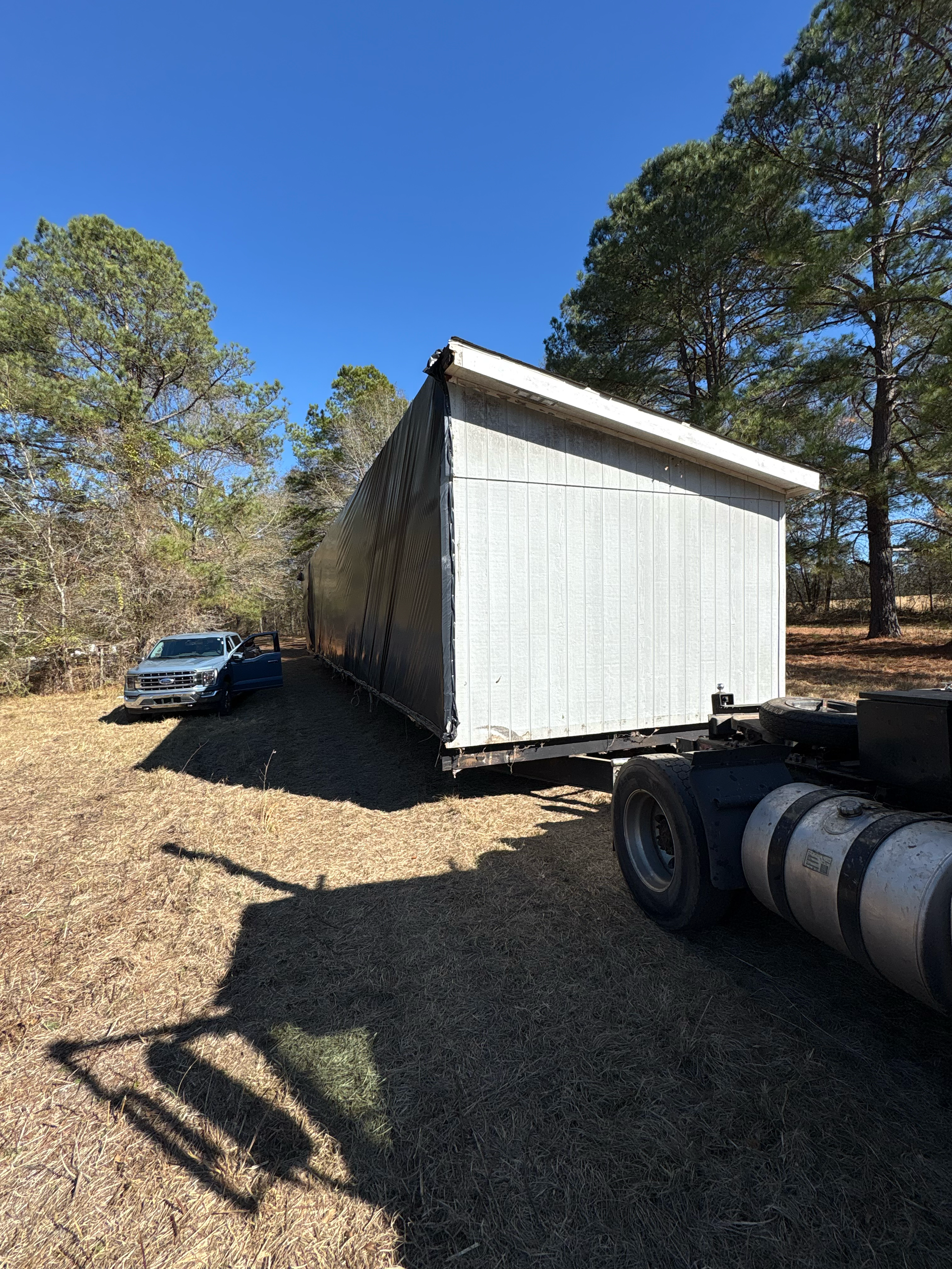A truck transports a long, white modular building on a trailer through a wooded area on a sunny day.