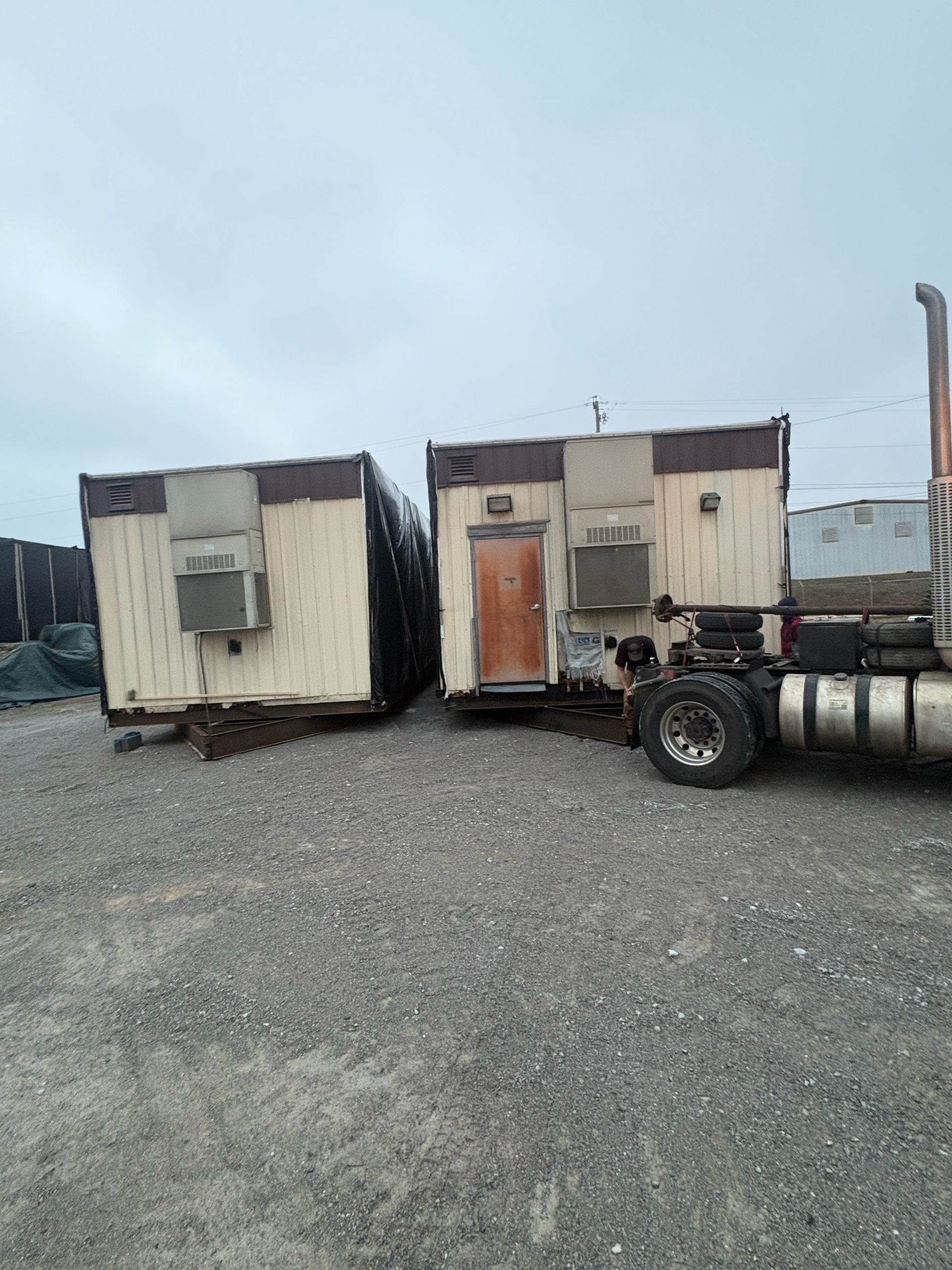 Two weathered portable office trailers with air conditioning units parked on a gravel lot next to a semi-truck.