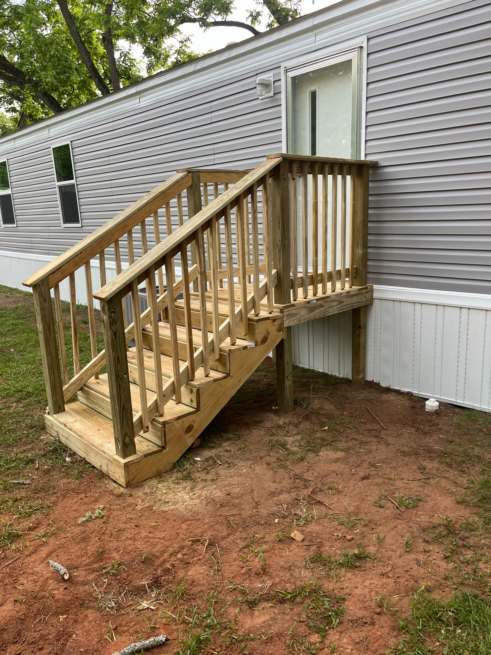 A wooden staircase with railings stands against the side of a light gray manufactured home with a white door.