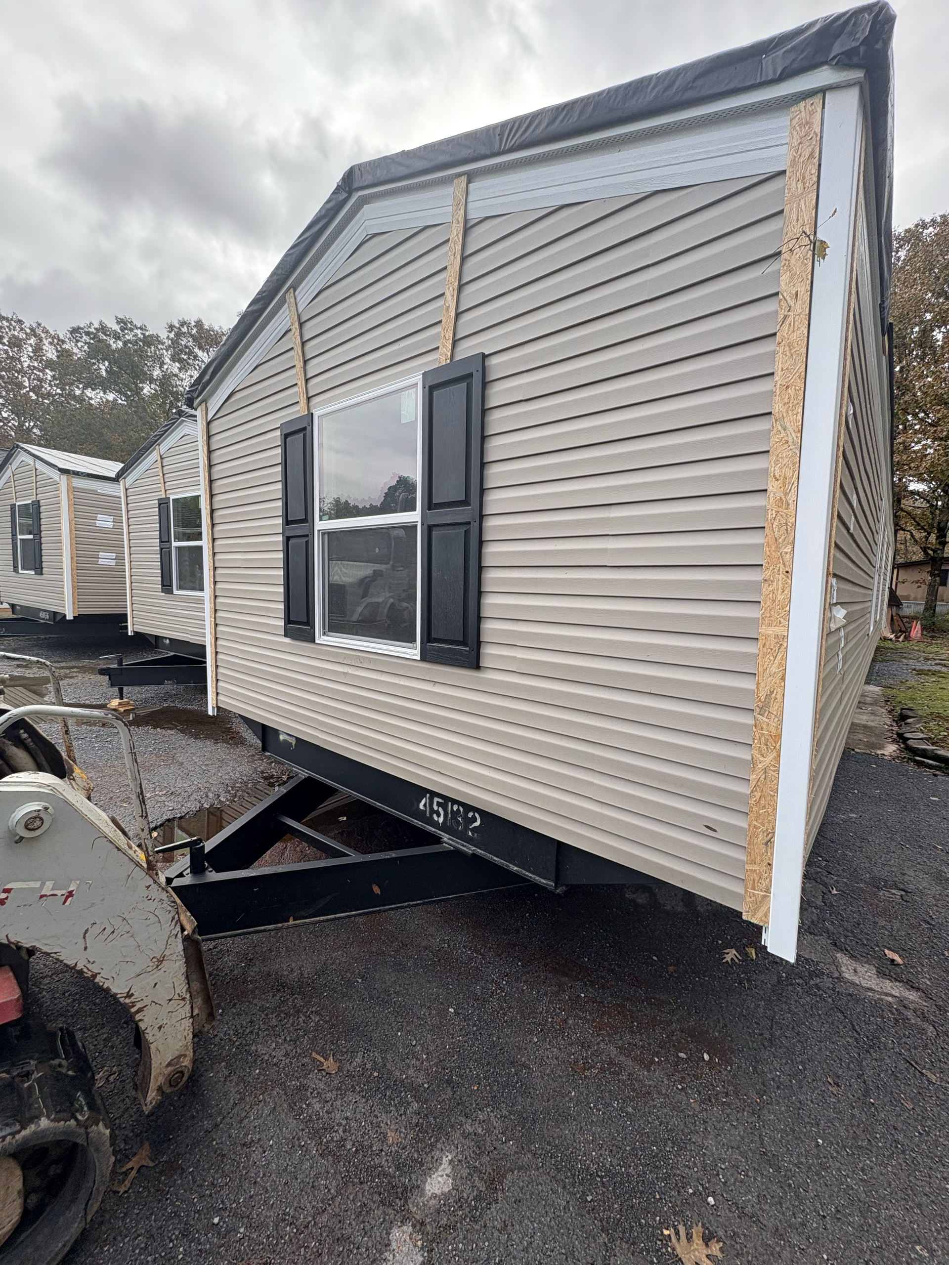 Tan, vinyl-sided mobile home unit on a trailer frame, parked outdoors on a gravel lot next to construction equipment.