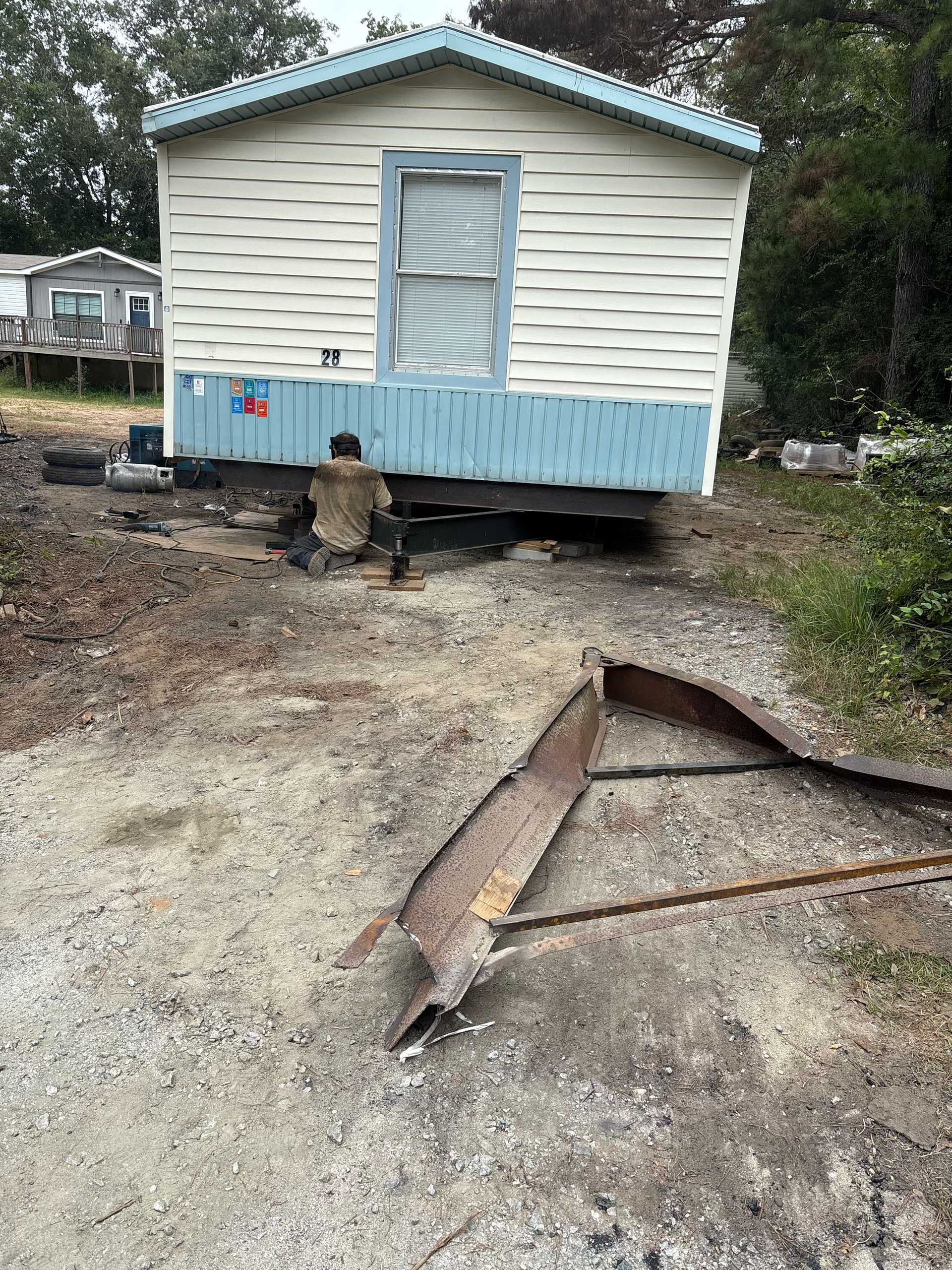 A person works beneath a light-colored mobile home, with a detached metal tow hitch lying on the gravel ground in front.