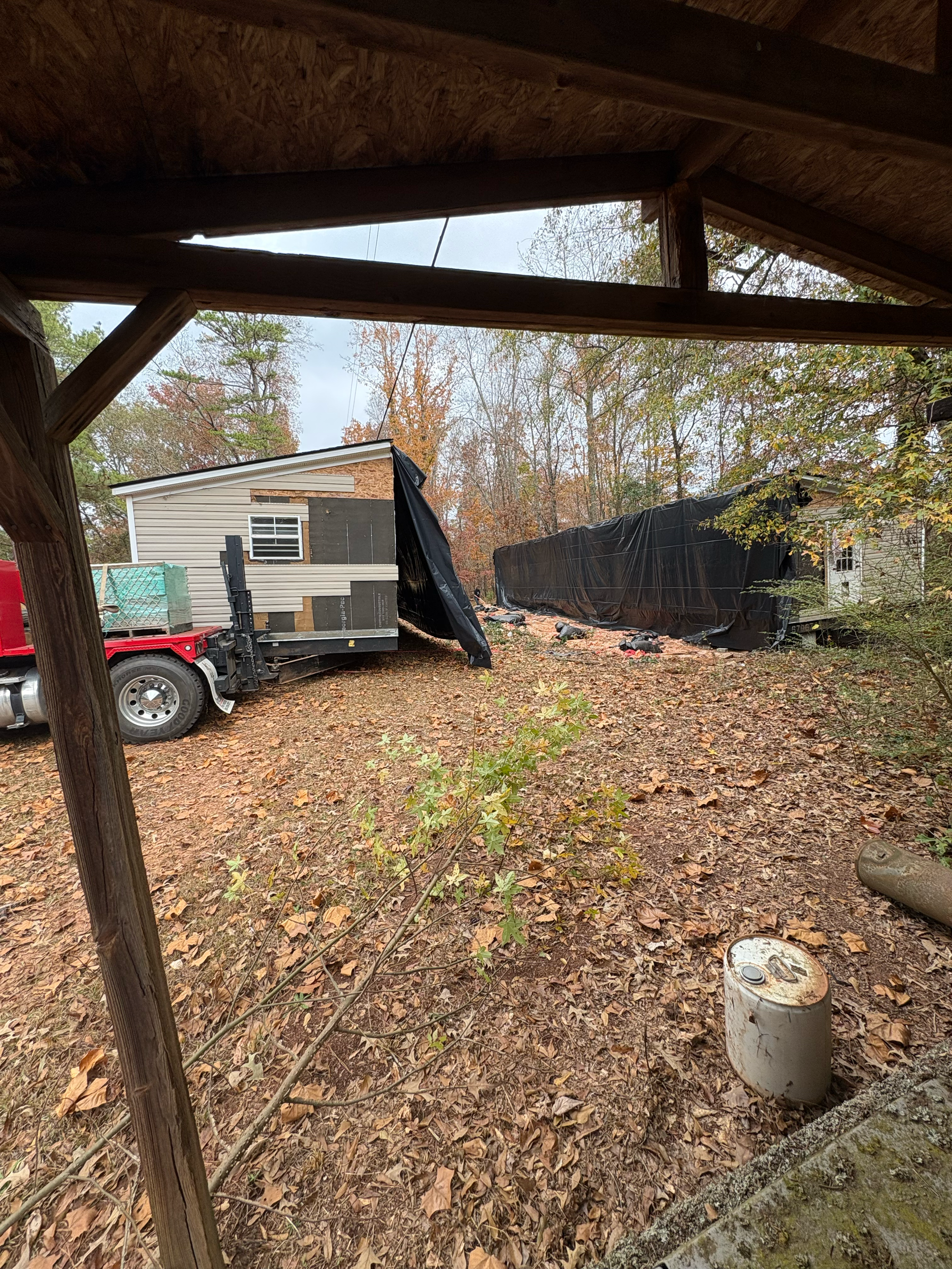 A red truck parked in a wooded area near a small, light-colored trailer and a long, dark structure covered by a tarp.