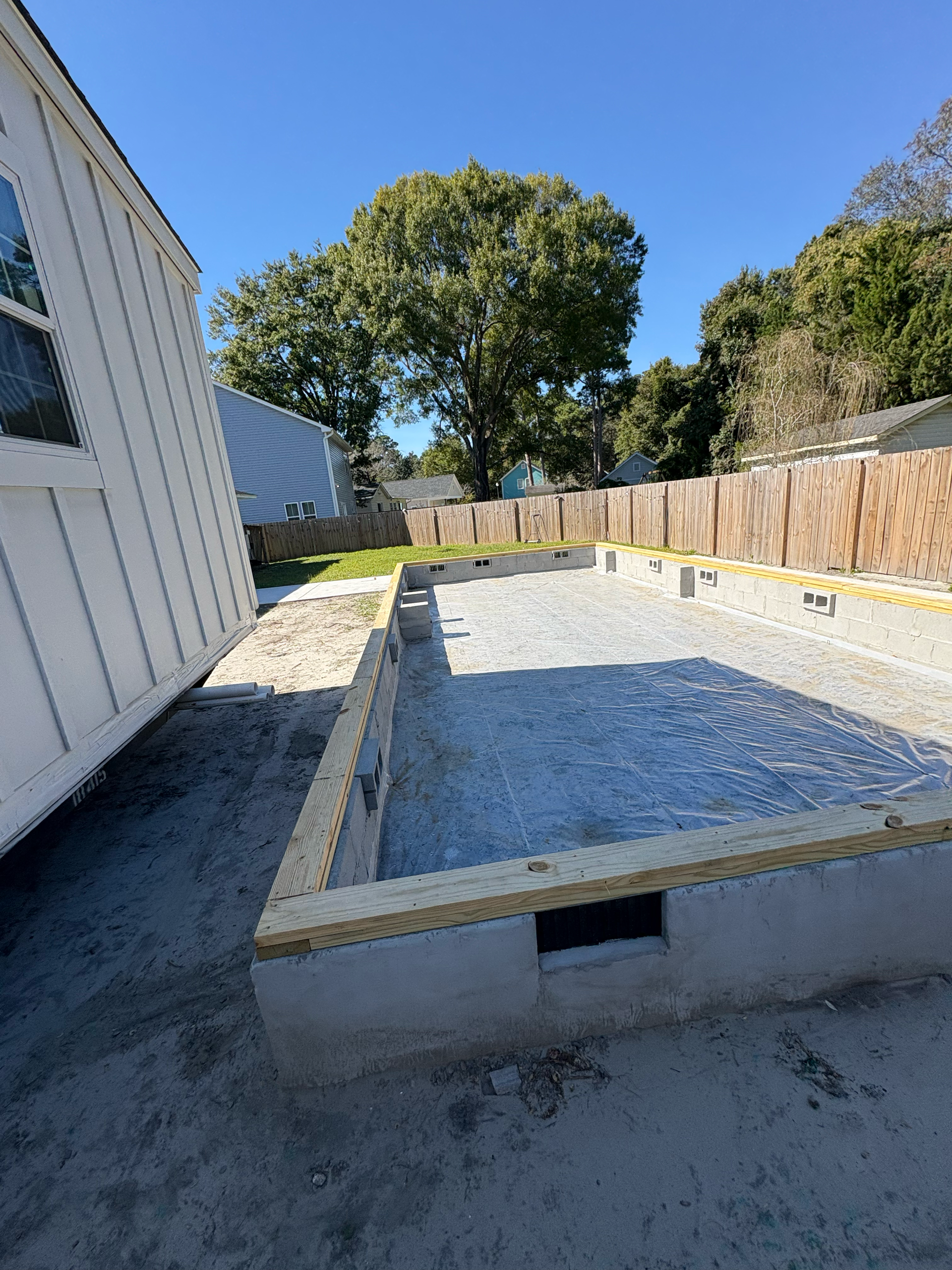 A rectangular concrete foundation with a gravel interior, topped by a wooden sill plate, adjacent to a white building.