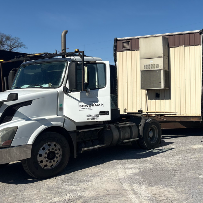 A white semi-truck parked outdoors, hooked to a tan, corrugated metal modular structure with an external HVAC unit.