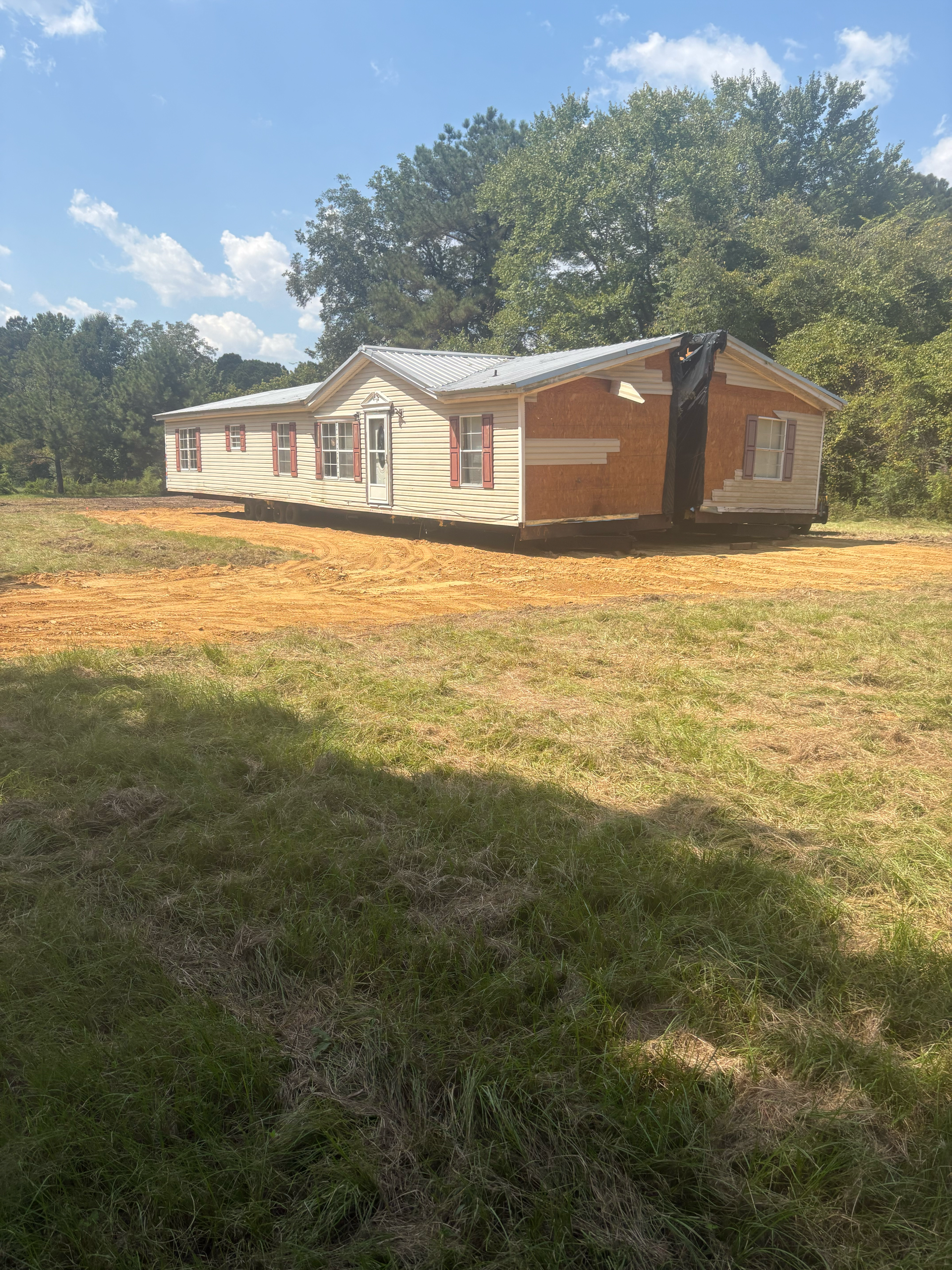 A single-story manufactured home sits on a dirt foundation in a grassy field surrounded by trees under a blue sky.