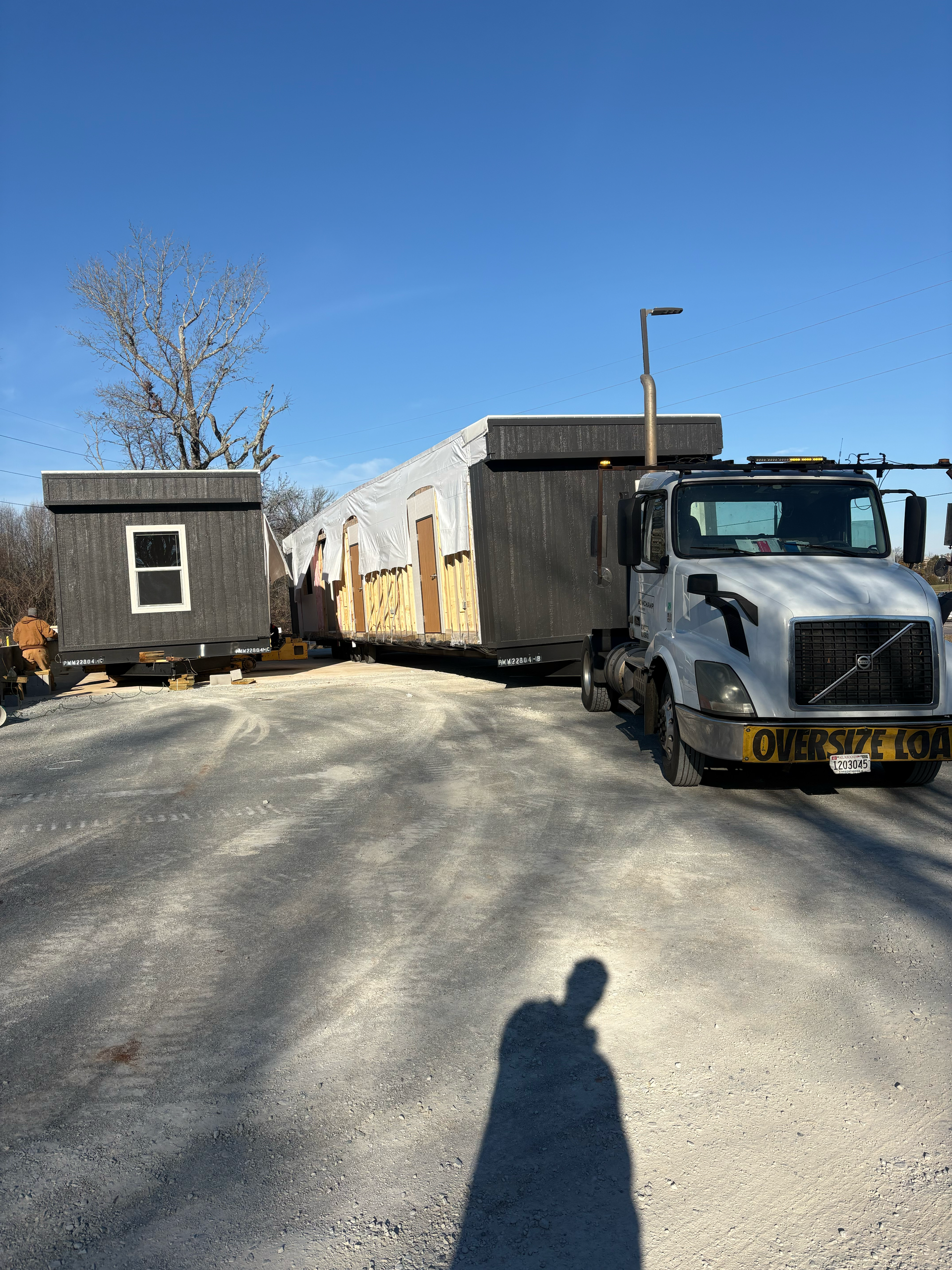 A semi-truck parked on a gravel lot next to two sections of a dark-sided modular home being transported.