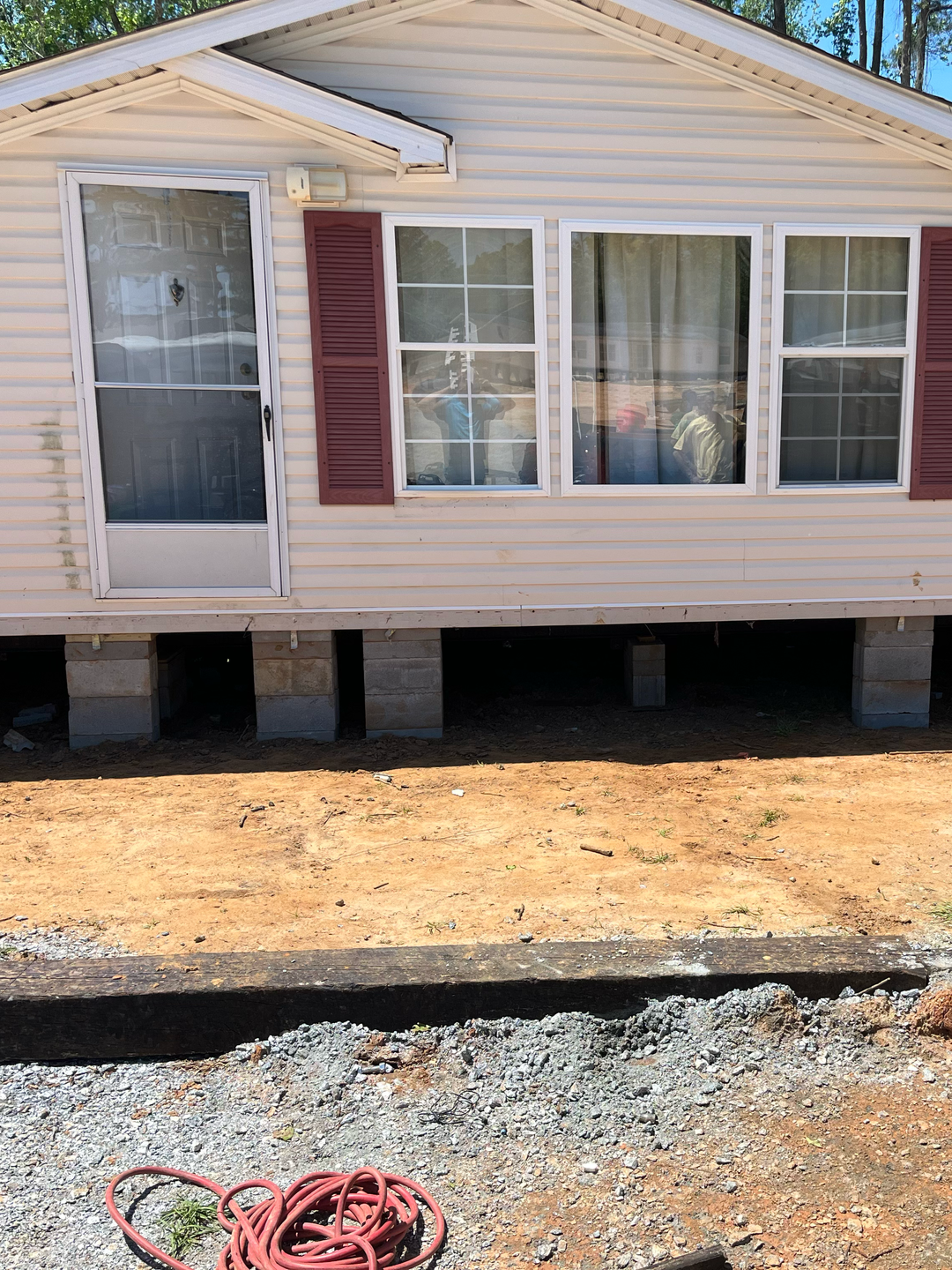 A tan mobile home with red shutters and a white storm door, elevated on concrete block piers above a dirt yard.